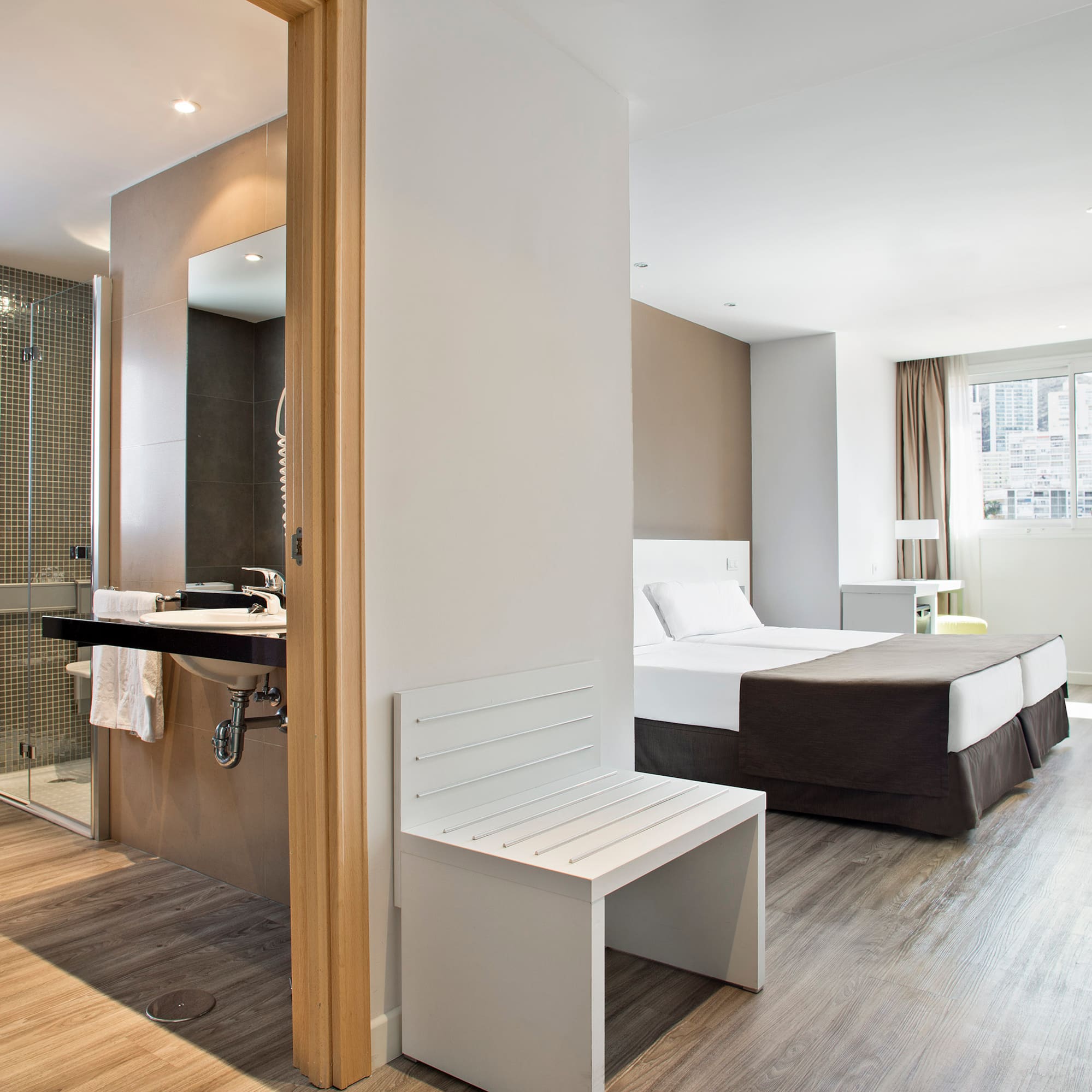 Modern hotel suite: twin beds, open bathroom, shower, and wood floor.