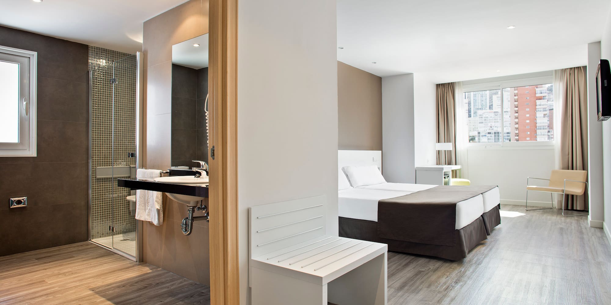 Modern hotel suite: twin beds, open bathroom, shower, and wood floor.