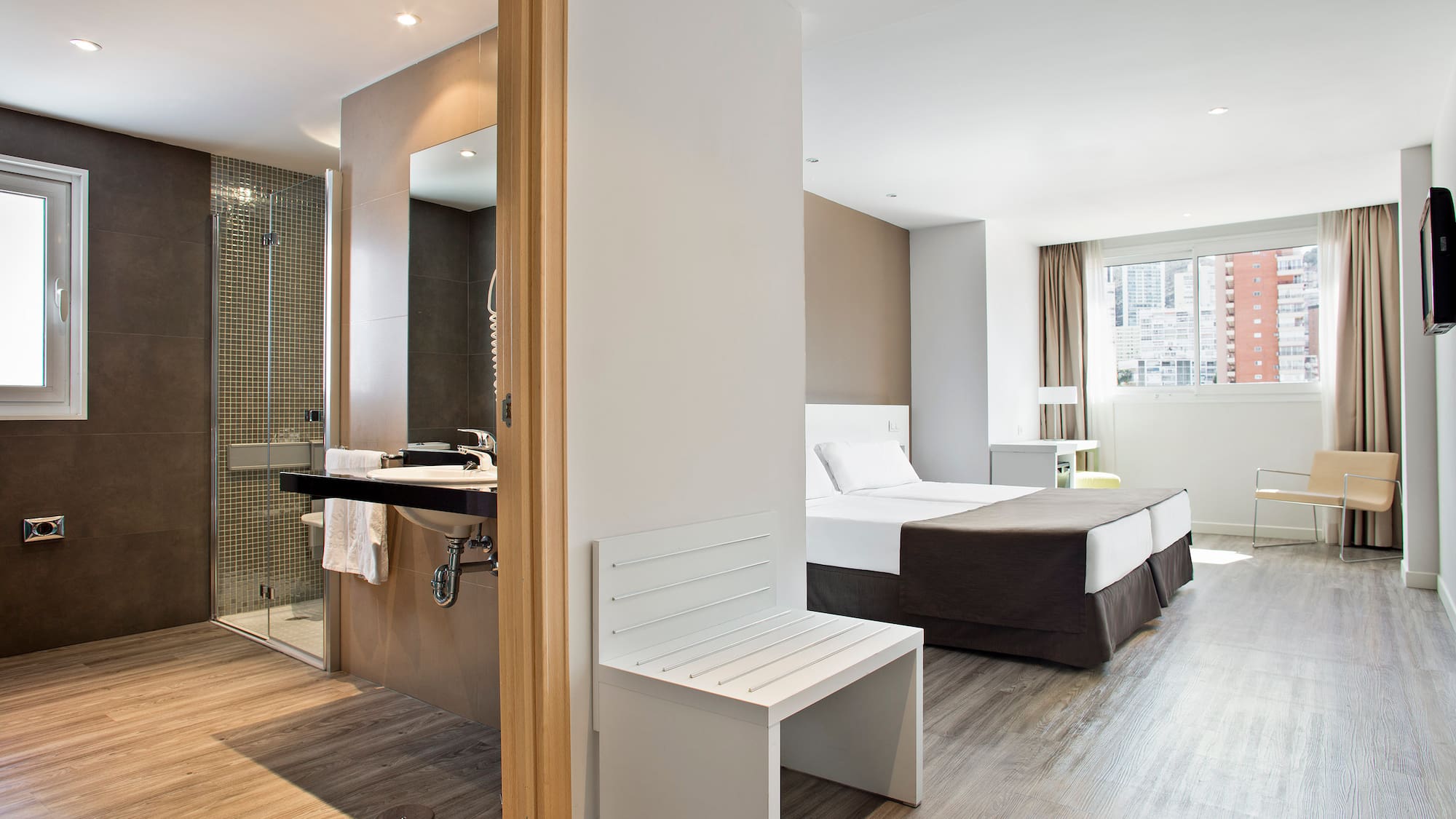 Modern hotel suite: twin beds, open bathroom, shower, and wood floor.