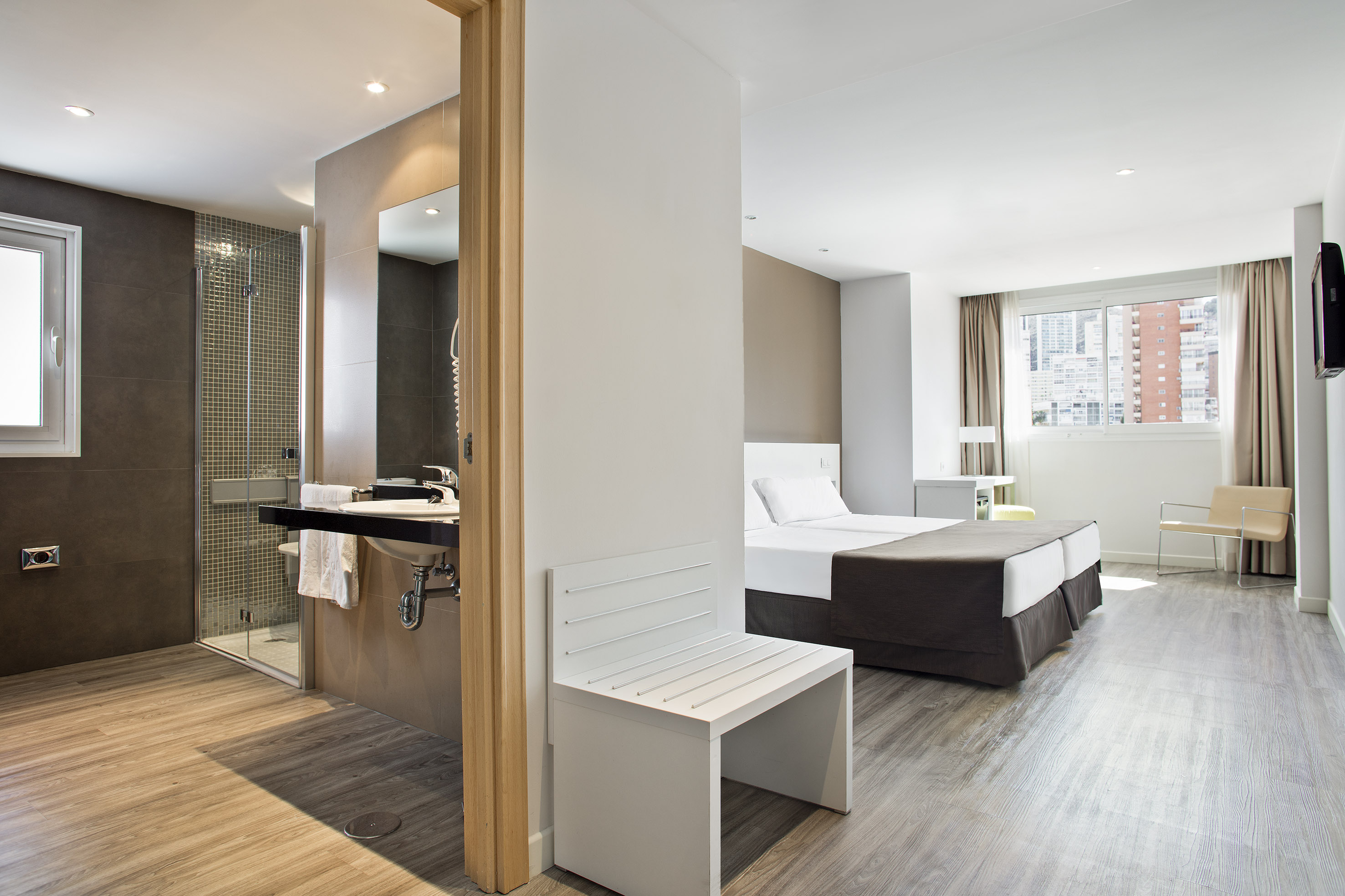 Modern hotel suite: twin beds, open bathroom, shower, and wood floor.