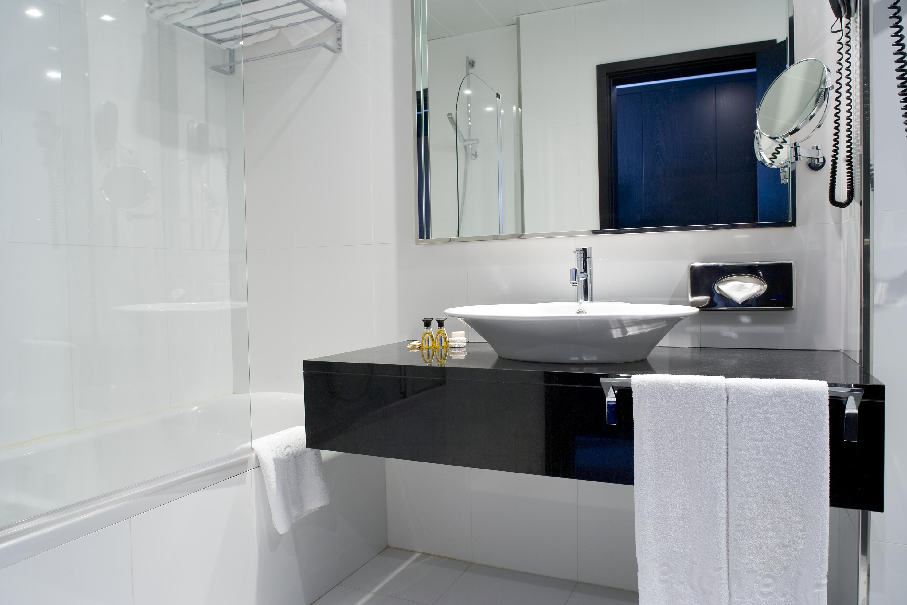 Modern hotel bathroom with white tiles, vessel sink, black counter, and bathtub.