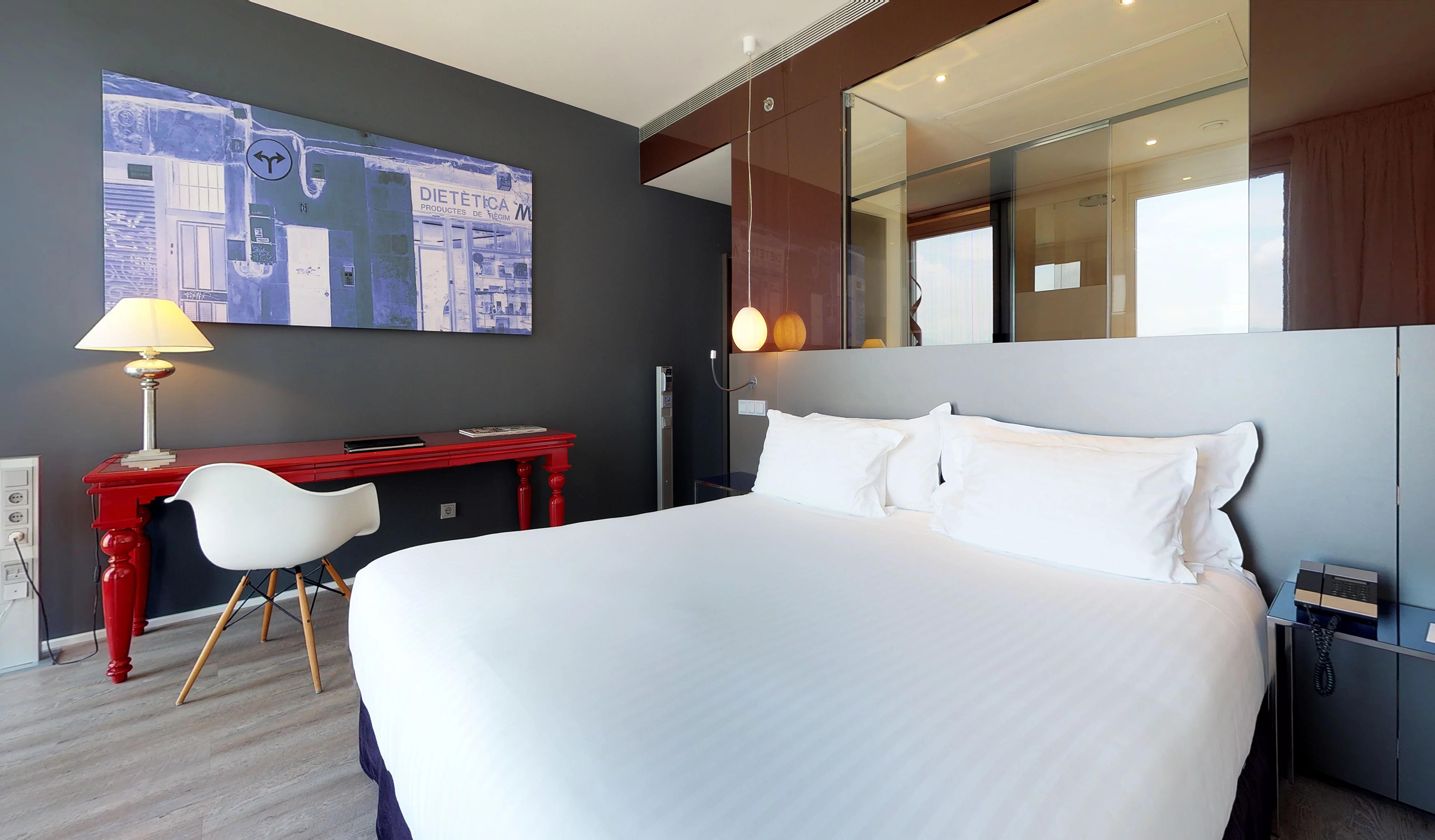 Modern hotel room with white bed, red desk, blue artwork, and glass partition.
