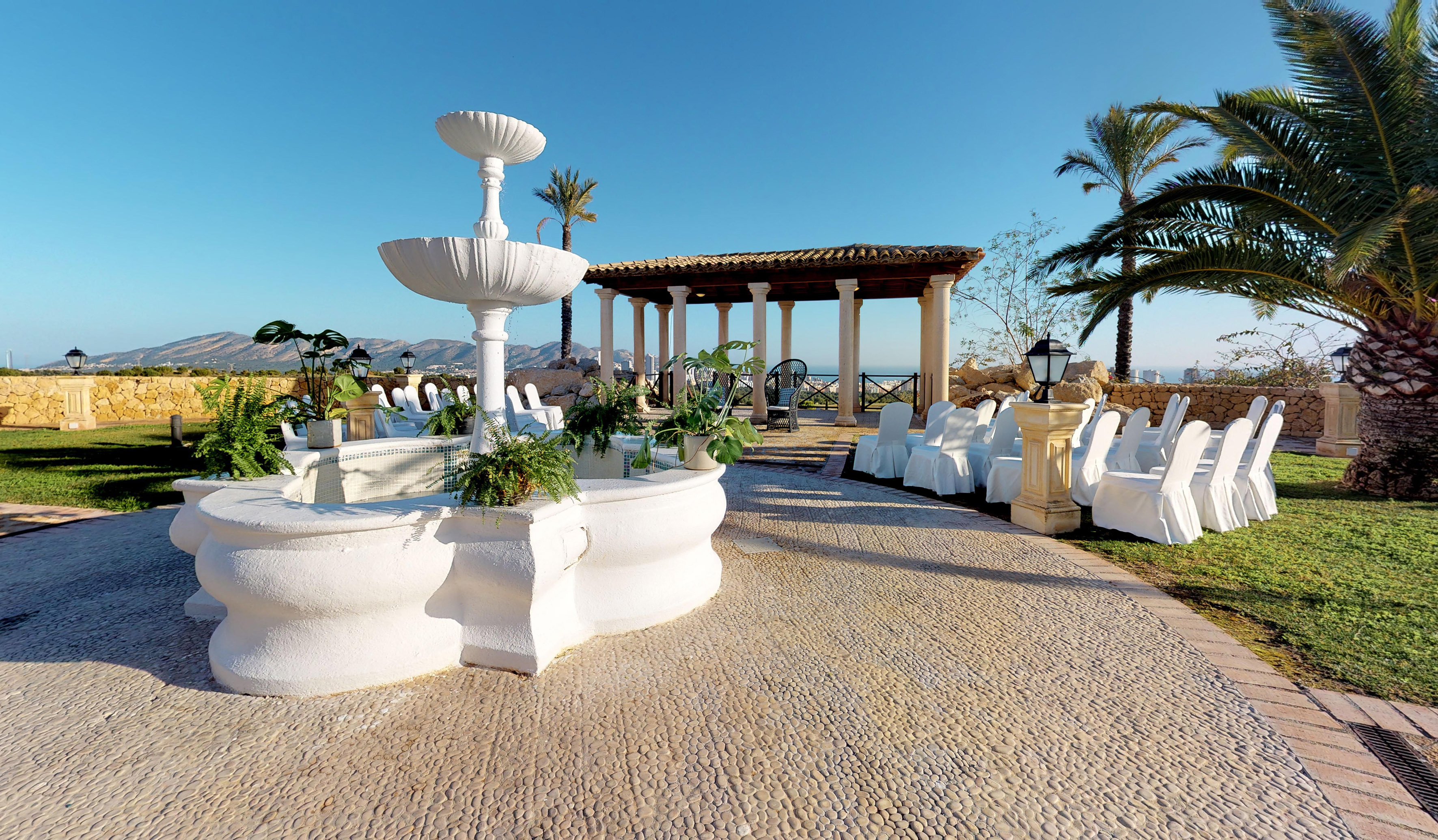 White fountain, pergola, and chairs at outdoor wedding venue overlooking sea.