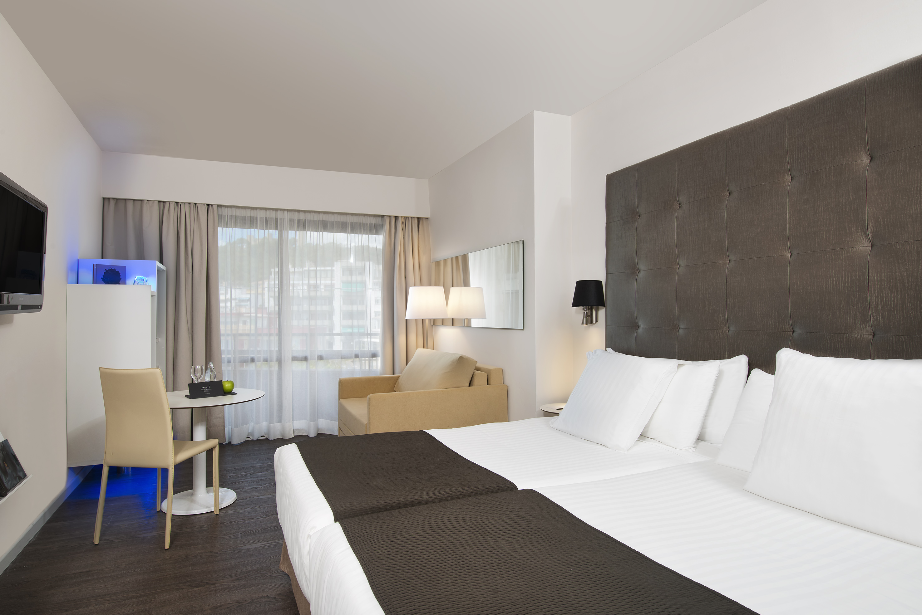 Modern hotel room with twin beds, dark headboard, sitting area, and large window.