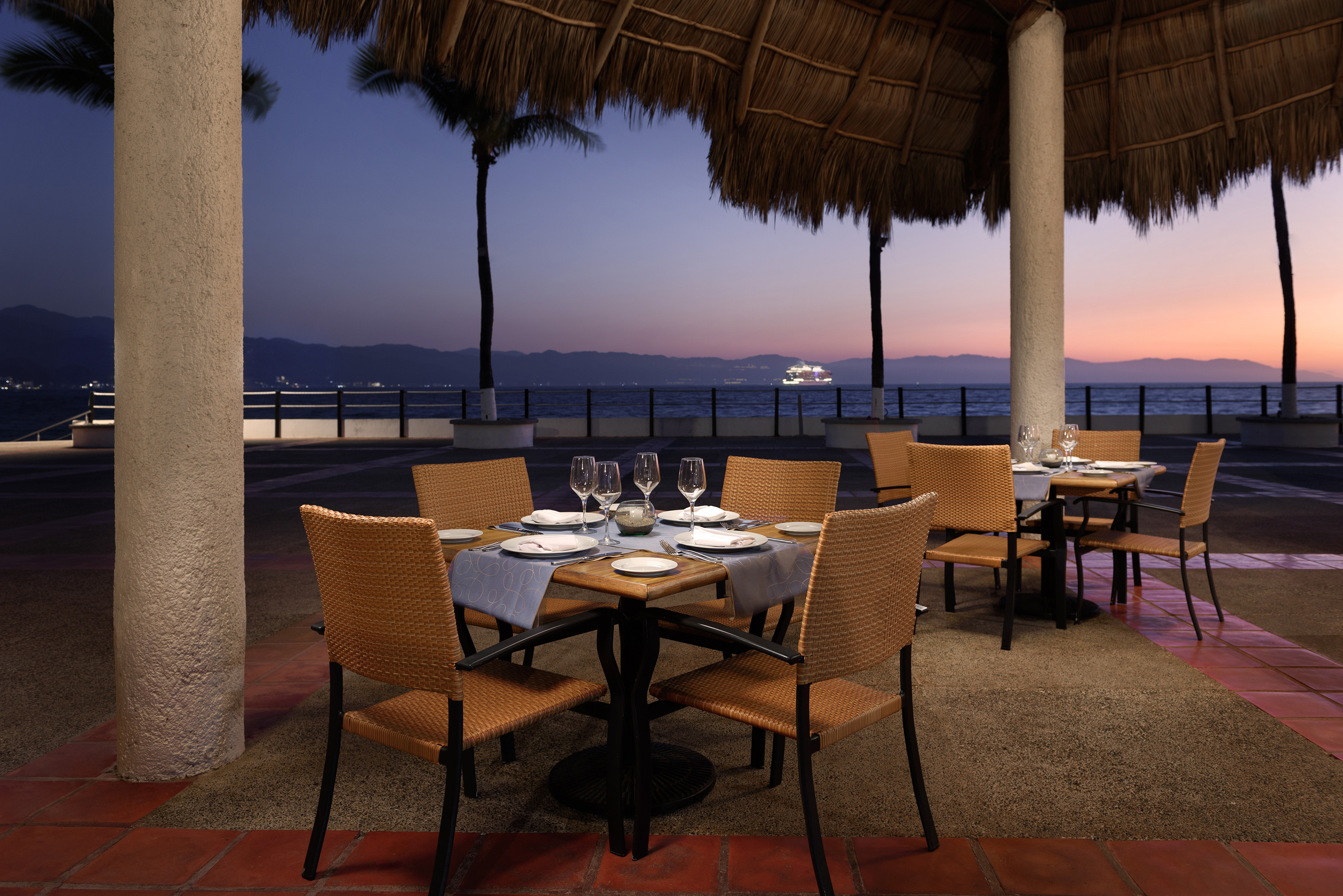 Tropical outdoor restaurant dining at sunset with ocean view and palm trees.