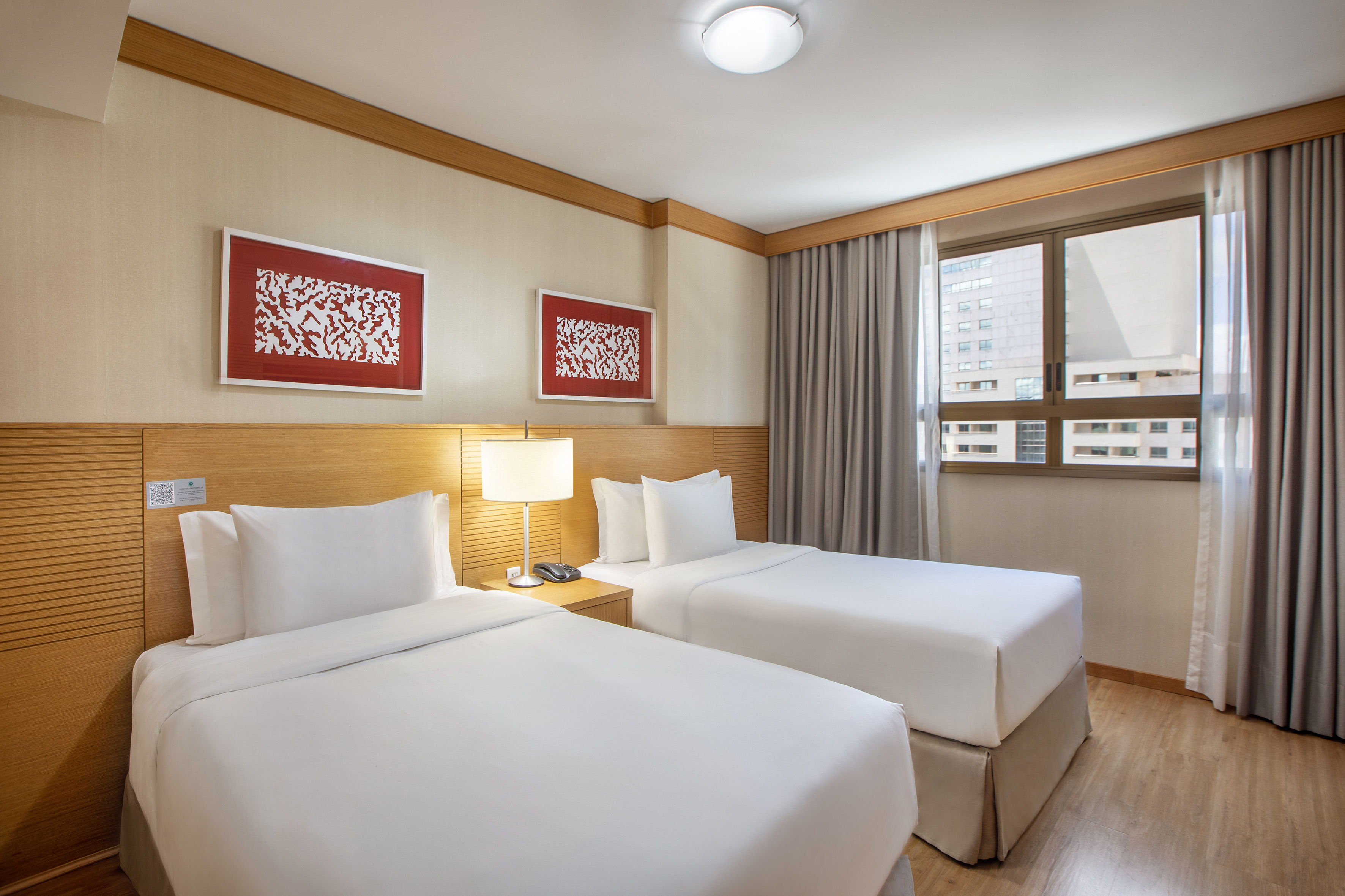 Hotel room with two twin beds, white linens, red abstract art, city window.