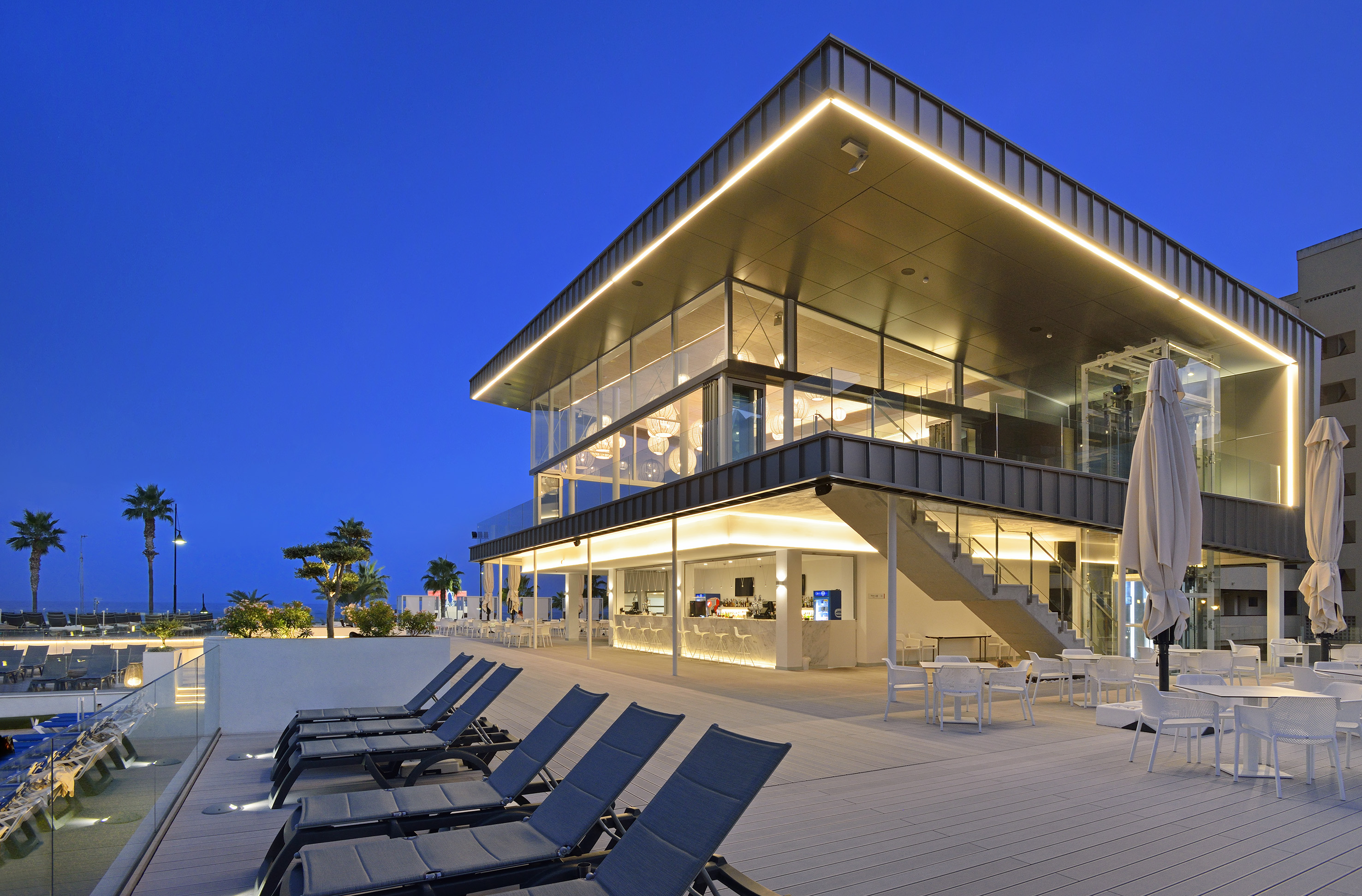 Modern illuminated resort bar and outdoor lounge terrace with palm trees.