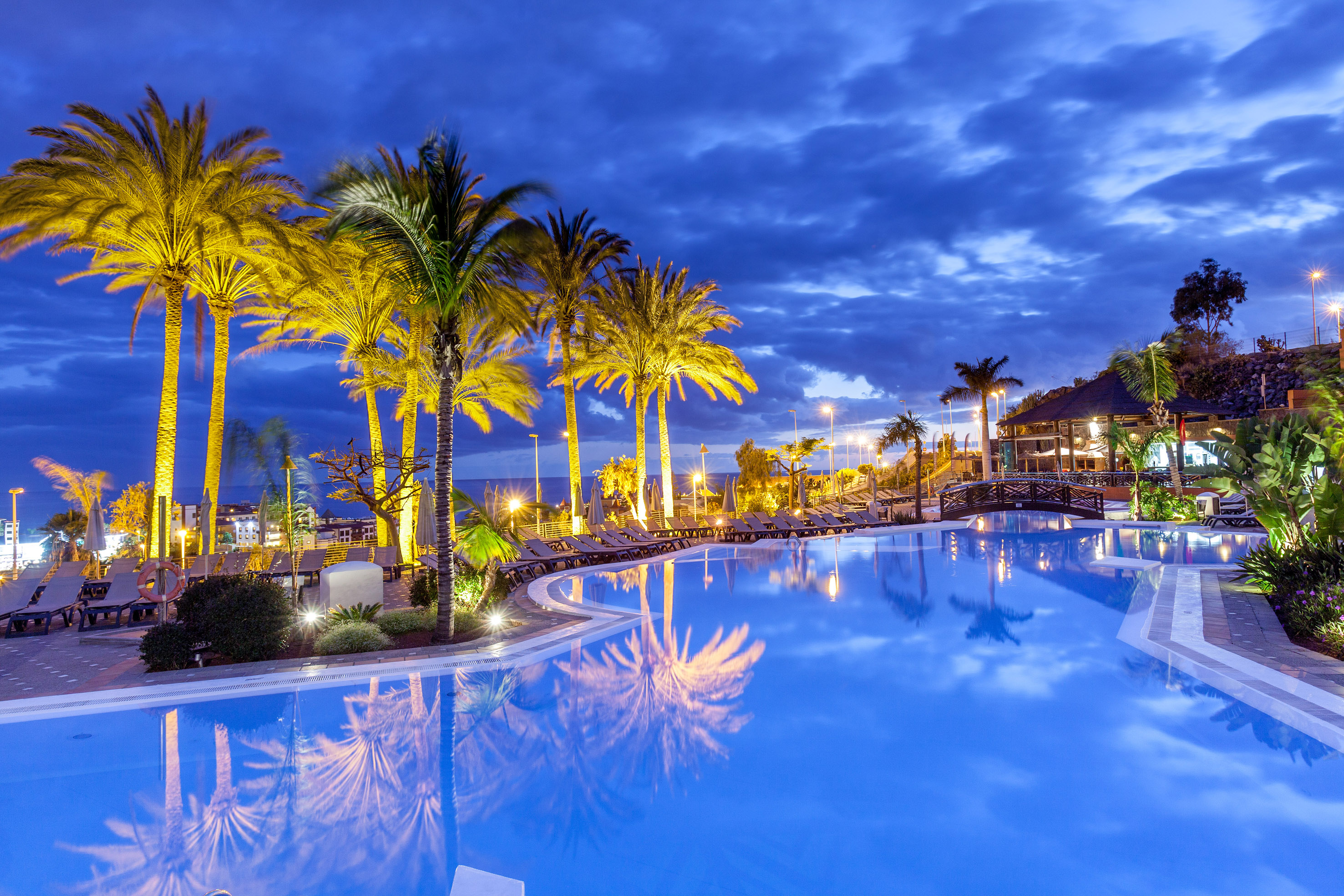 Illuminated tropical resort pool at night with palm trees reflecting in blue water.