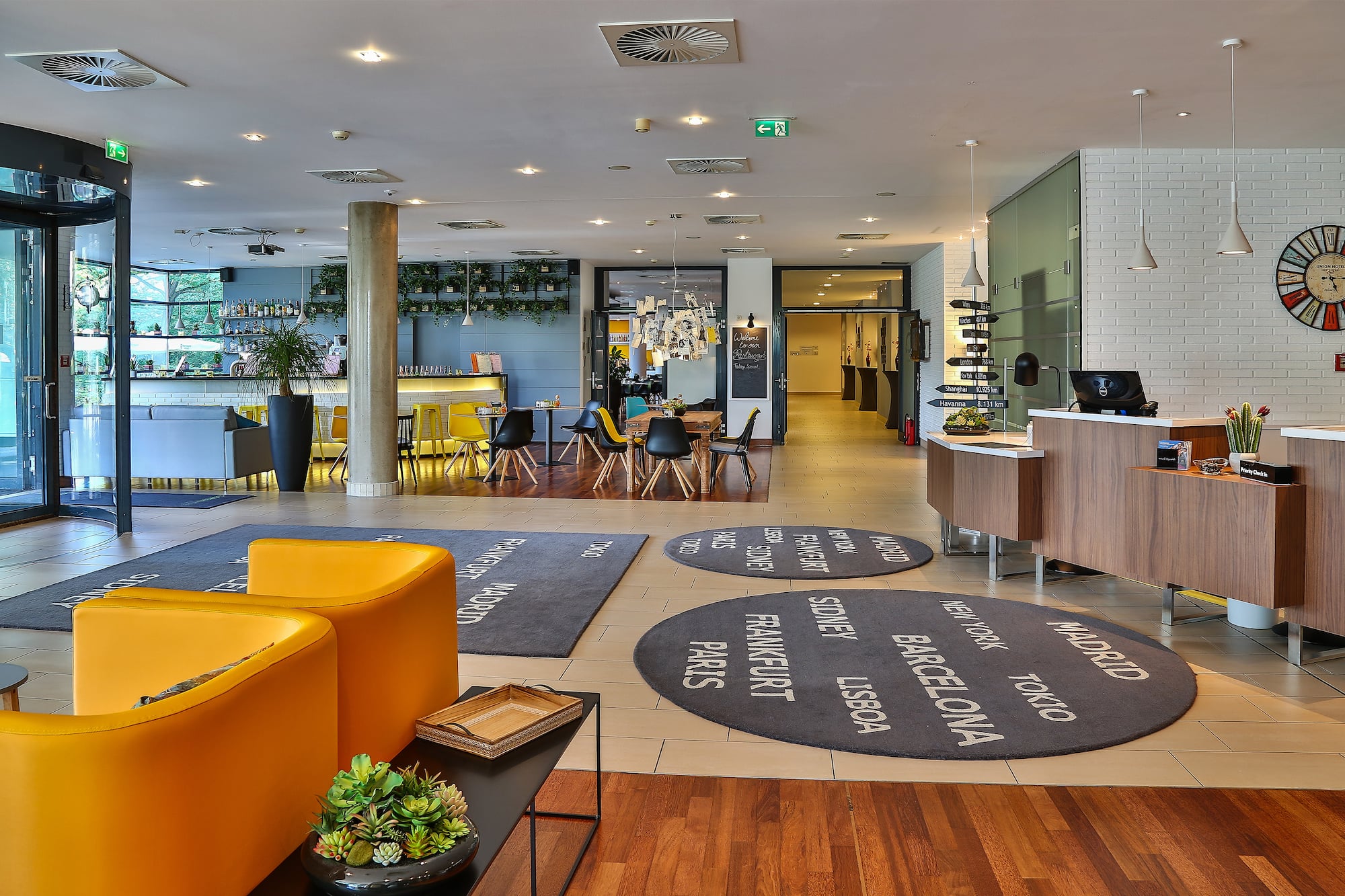 Modern hotel lobby with reception, bar, yellow lounge chairs, and city rugs.