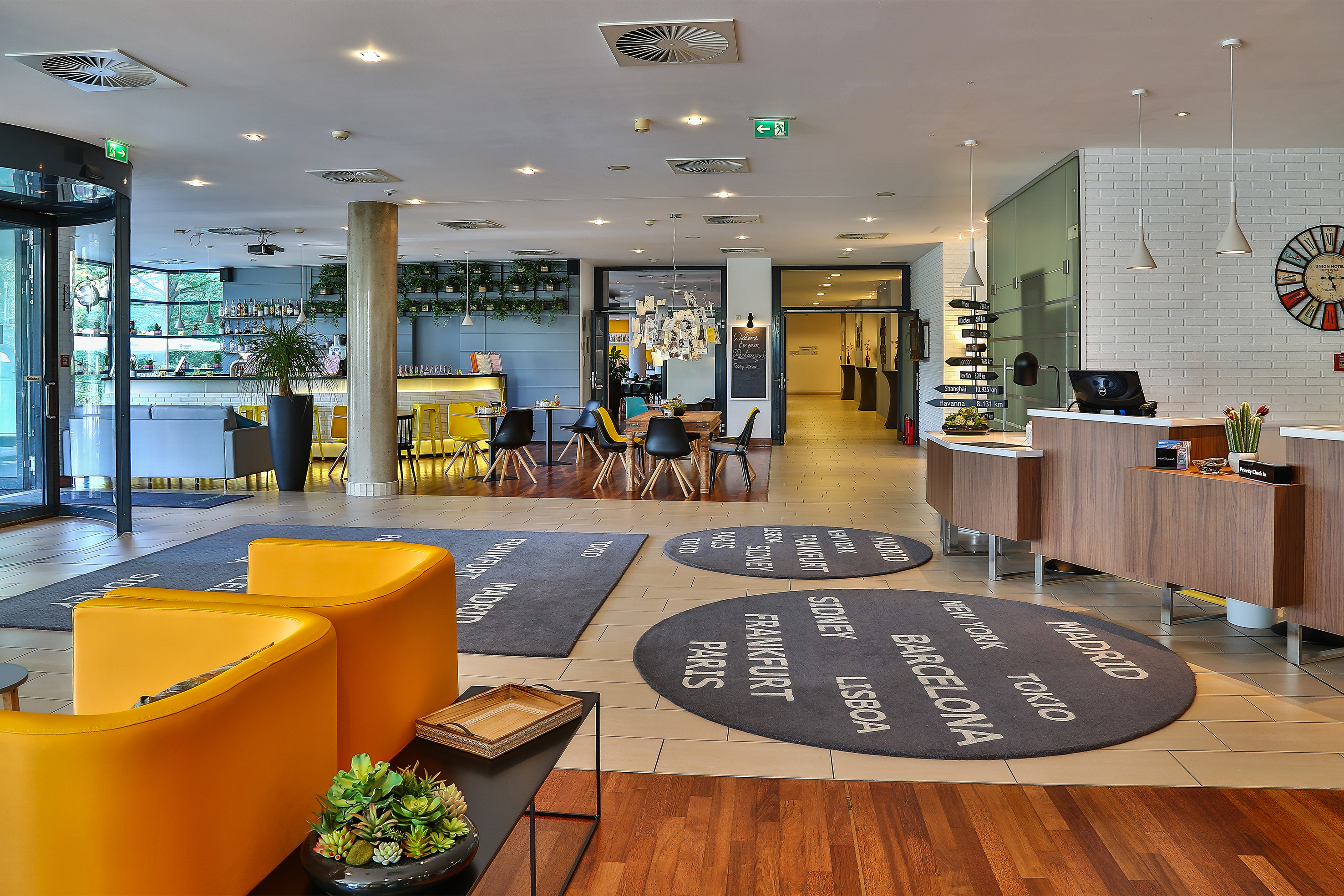 Modern hotel lobby with reception, bar, yellow lounge chairs, and city rugs.