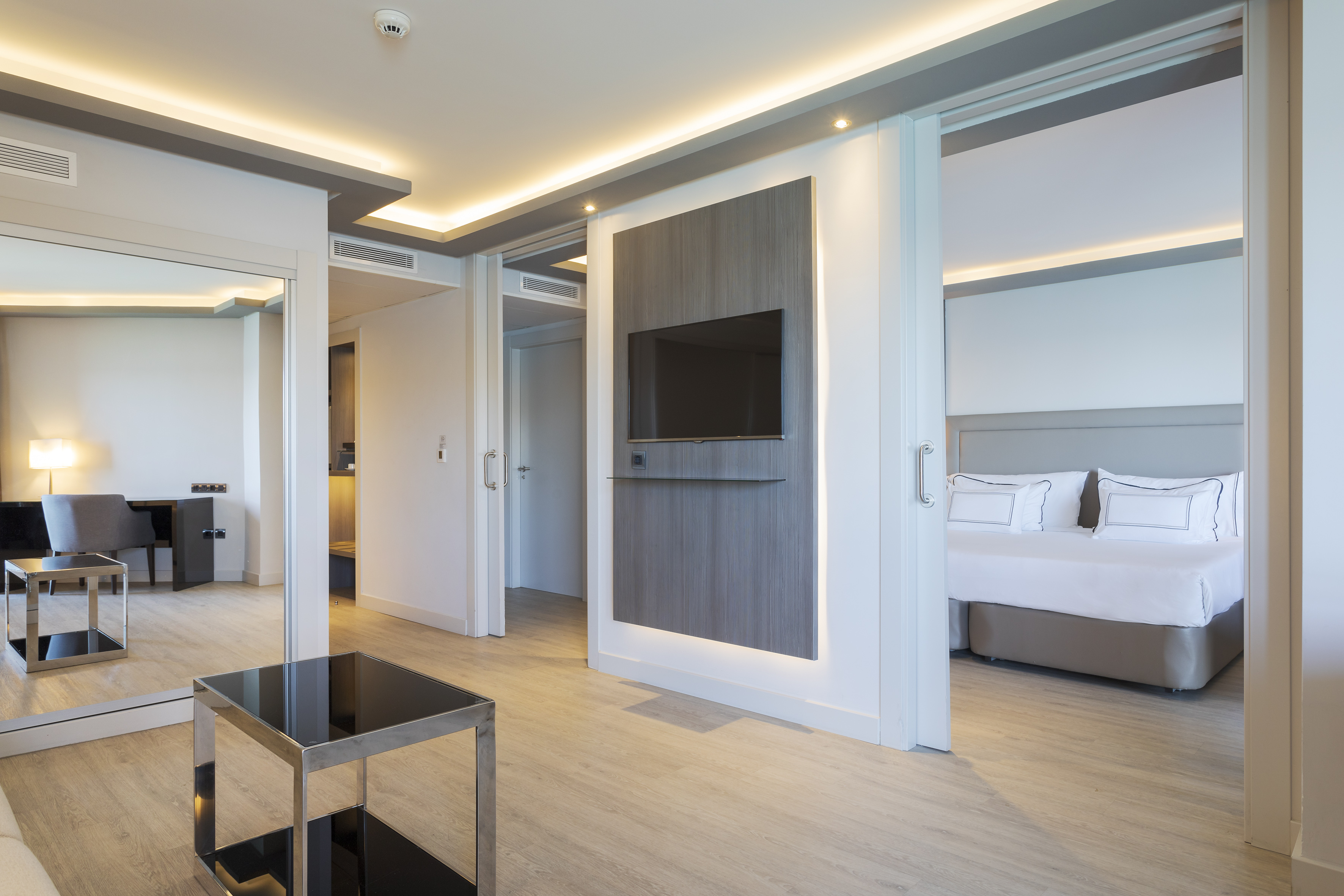 Modern hotel suite with living area, wall TV, and bedroom behind sliding door.
