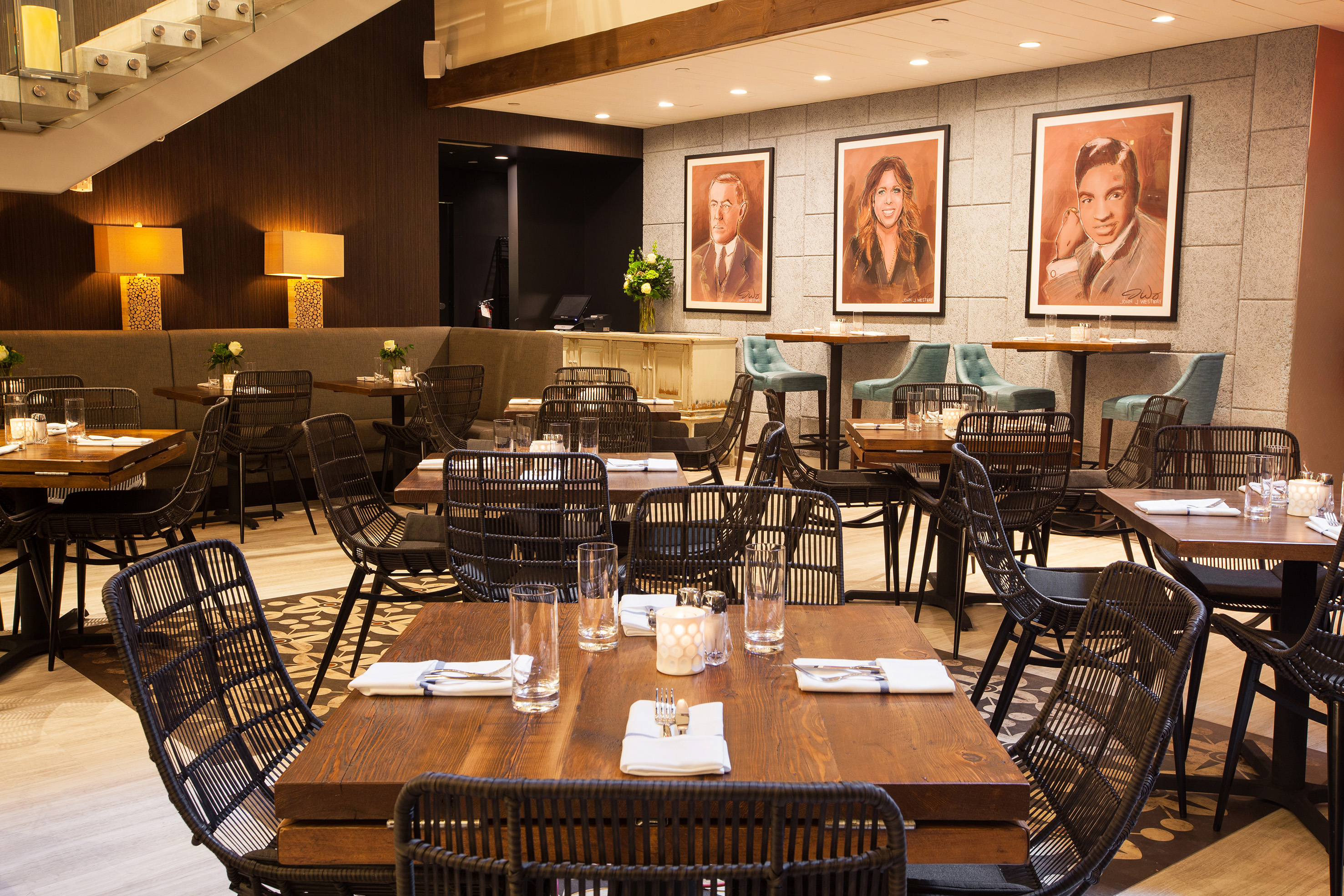 Modern restaurant dining room with wooden tables, black woven chairs, and framed portraits.