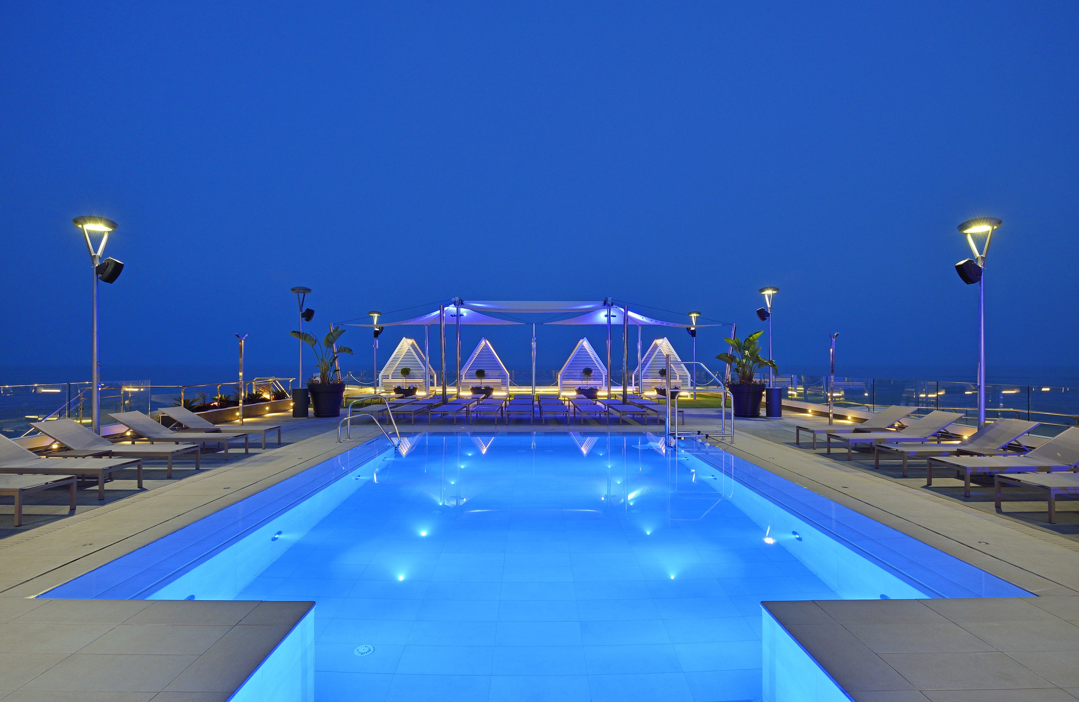 Blue lit rooftop pool at night with modern loungers and ocean views.