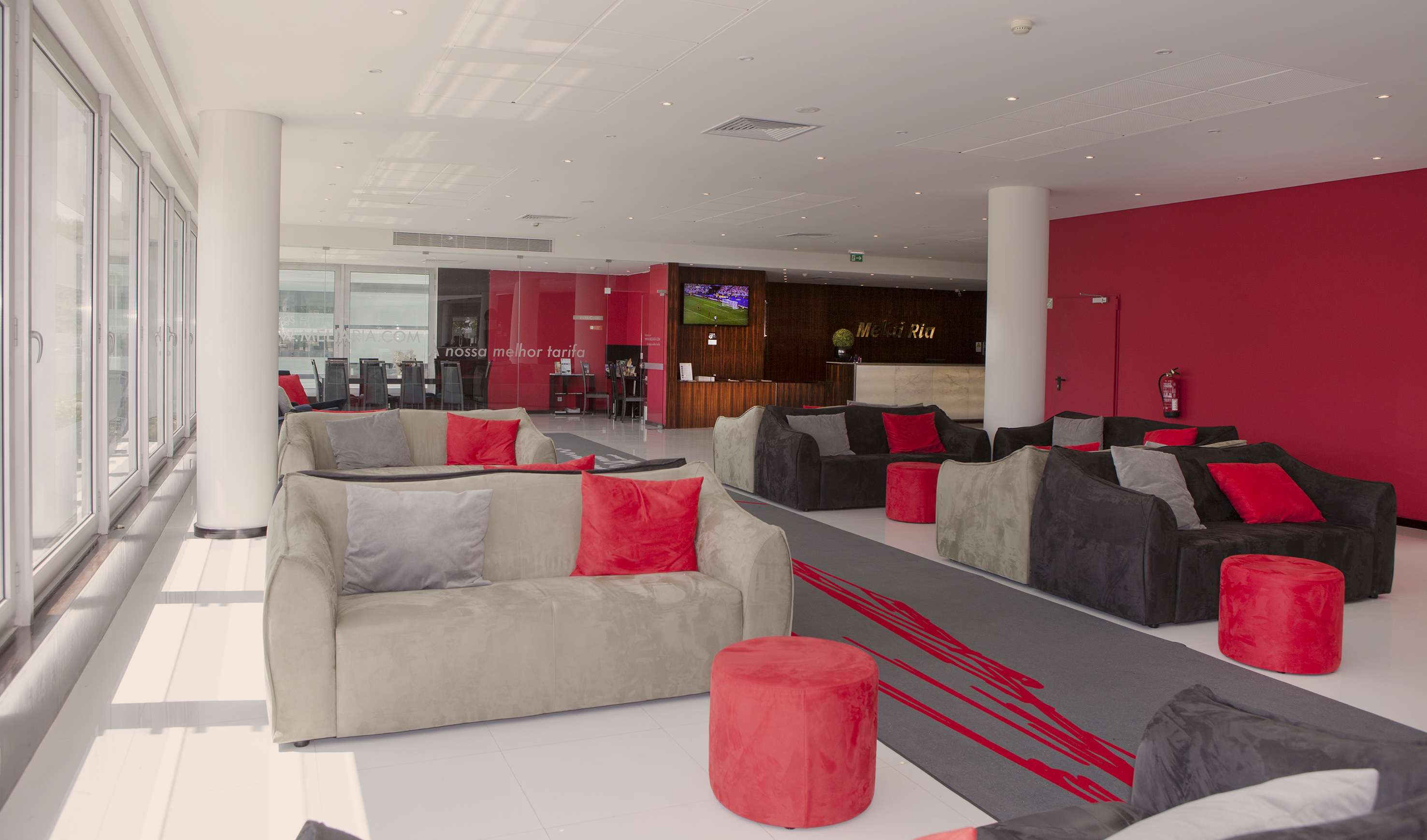 Modern hotel lobby with grey sofas, red cushions, reception area, and large windows.