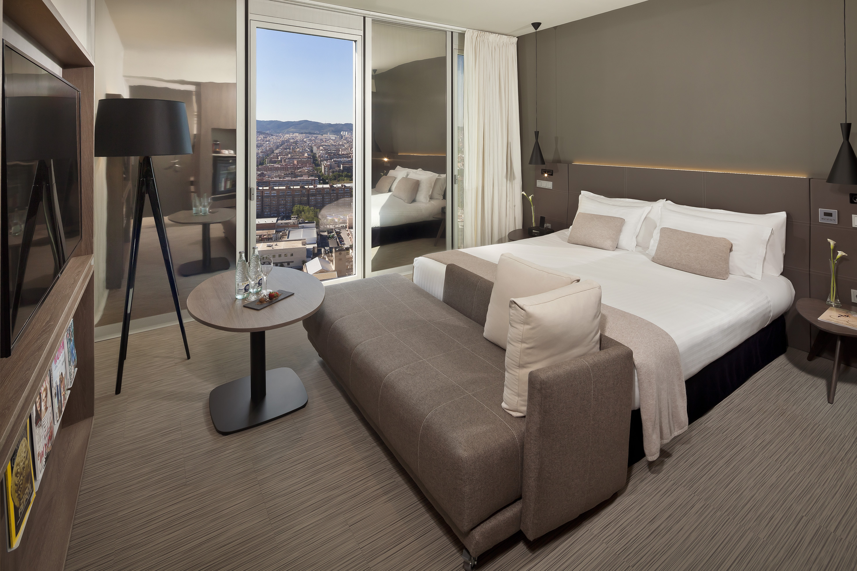 Modern hotel bedroom with a large bed, sofa, and panoramic city view.