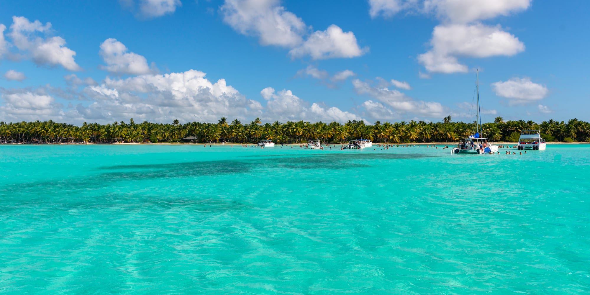 Aguas turquesas del Caribe, palmeras, barcos y bañistas en playa tropical.