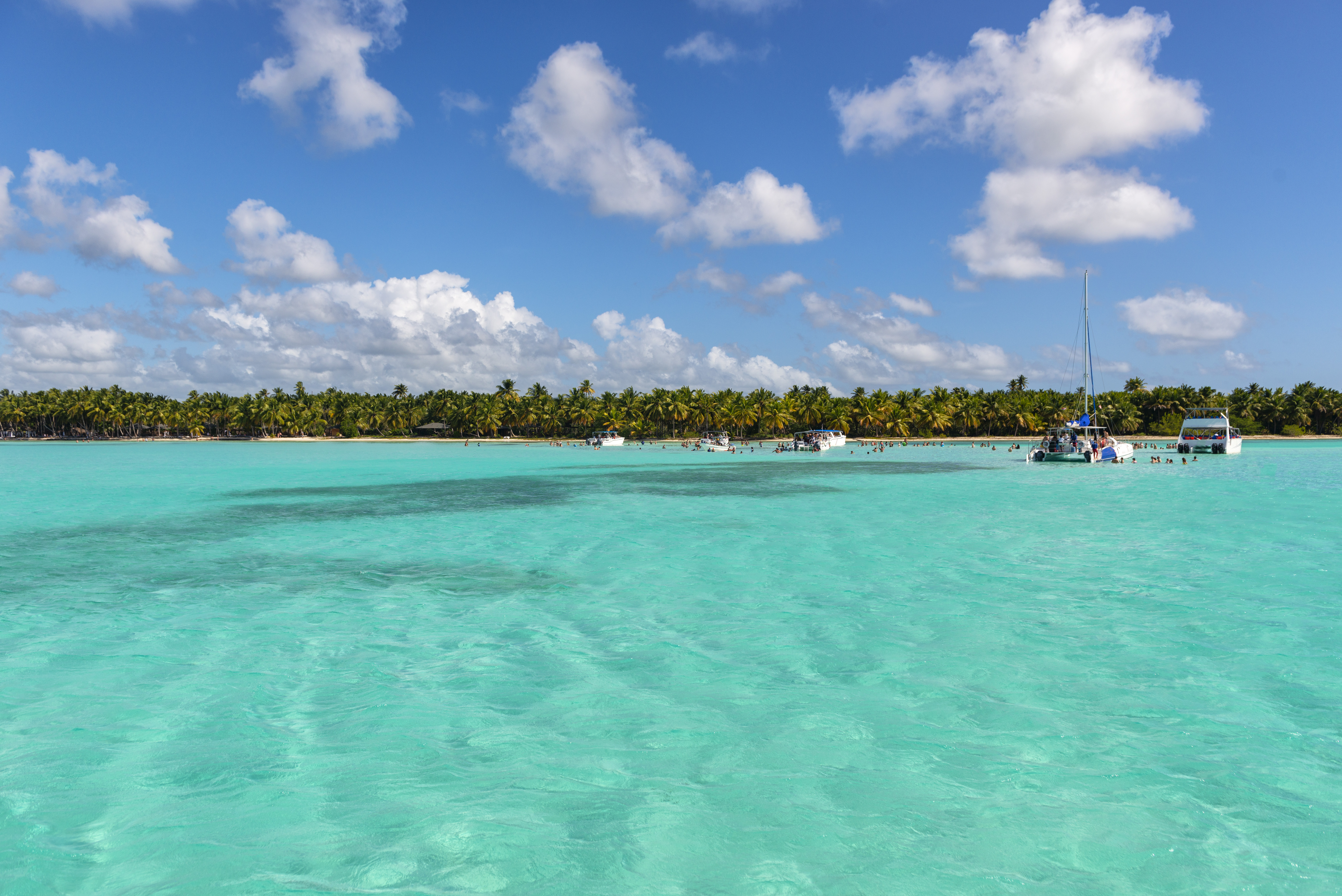 Agua turquesa Caribe, playa tropical con palmeras, barcos y gente en el mar.