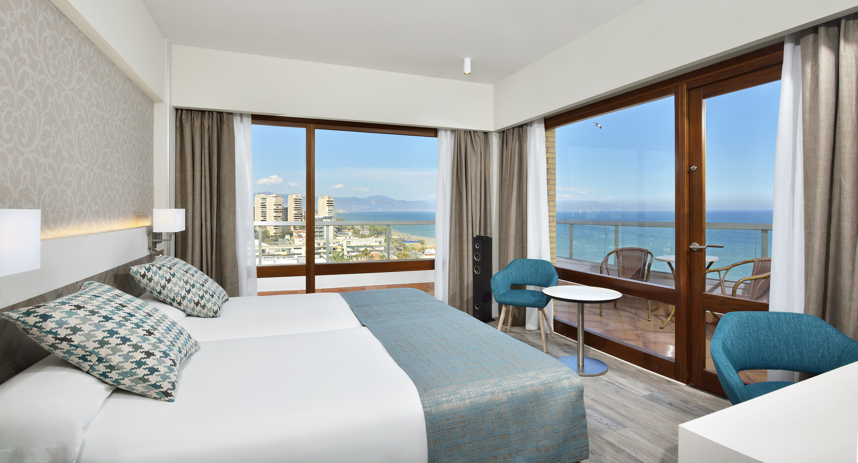 Bright hotel room with twin beds, sea view balcony, and city coastline.