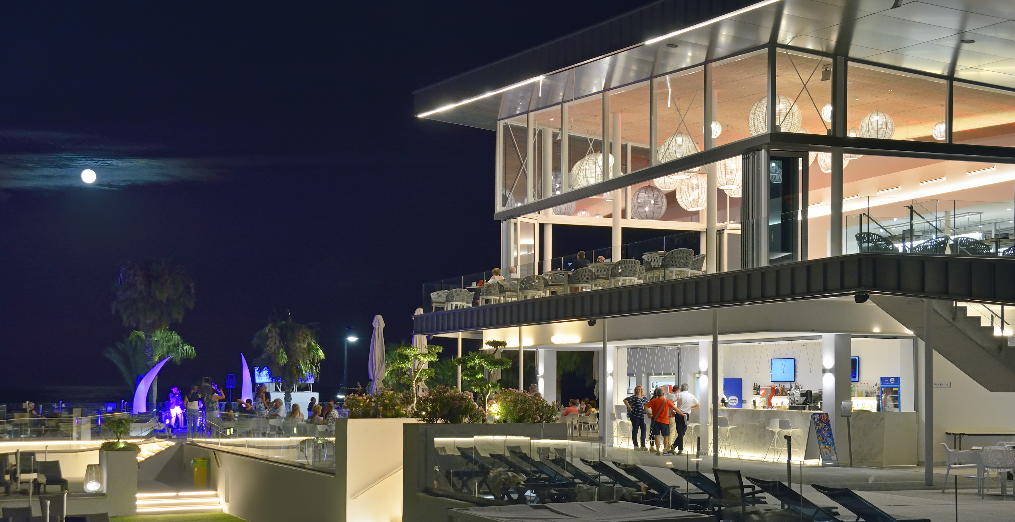 Modern illuminated resort restaurant & bar at night with outdoor seating, moon.
