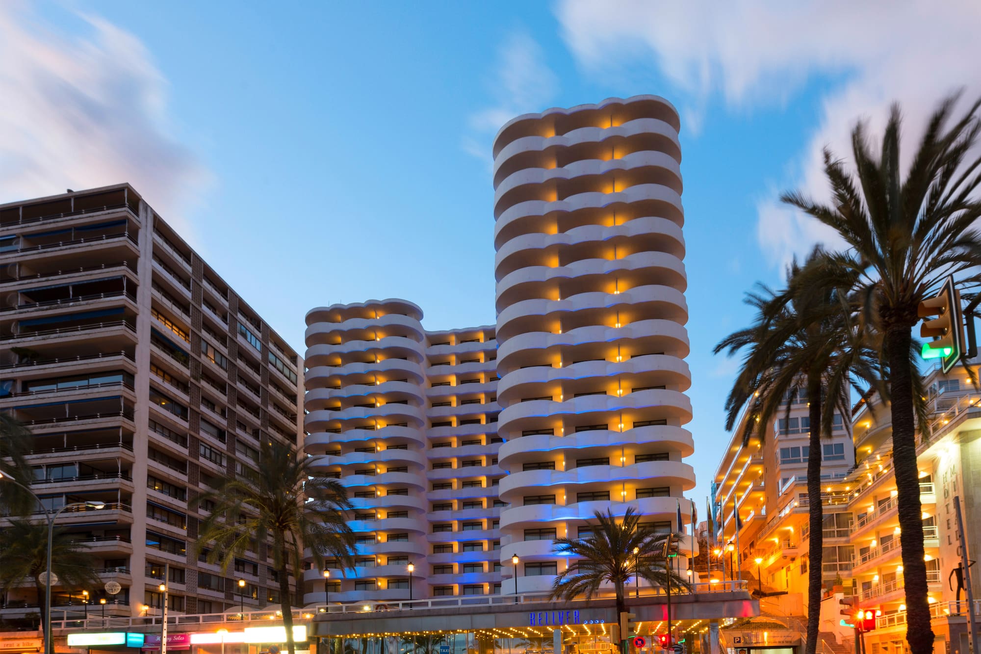 Illuminated Palma hotel with wavy balconies, palm trees, and city street at dusk.
