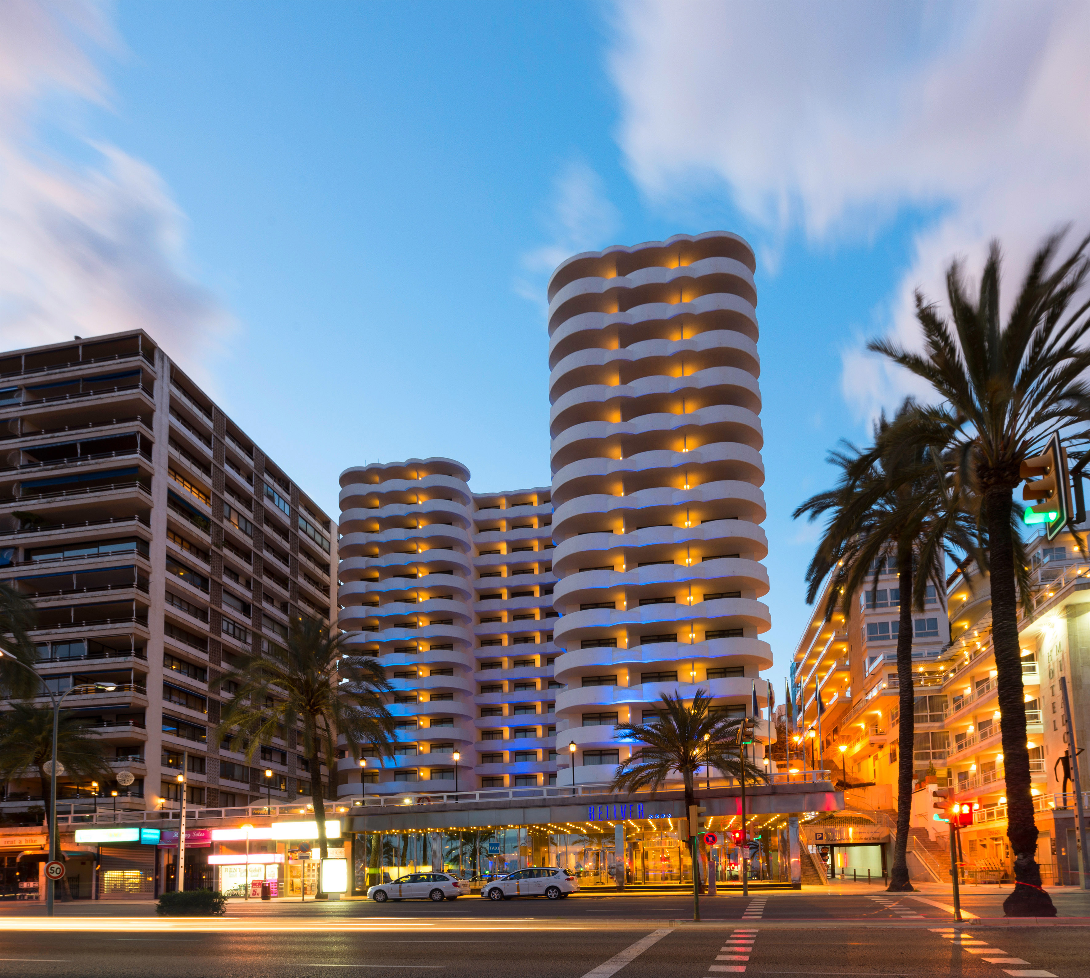 Illuminated Palma hotel with wavy balconies, palm trees, and city street at dusk.