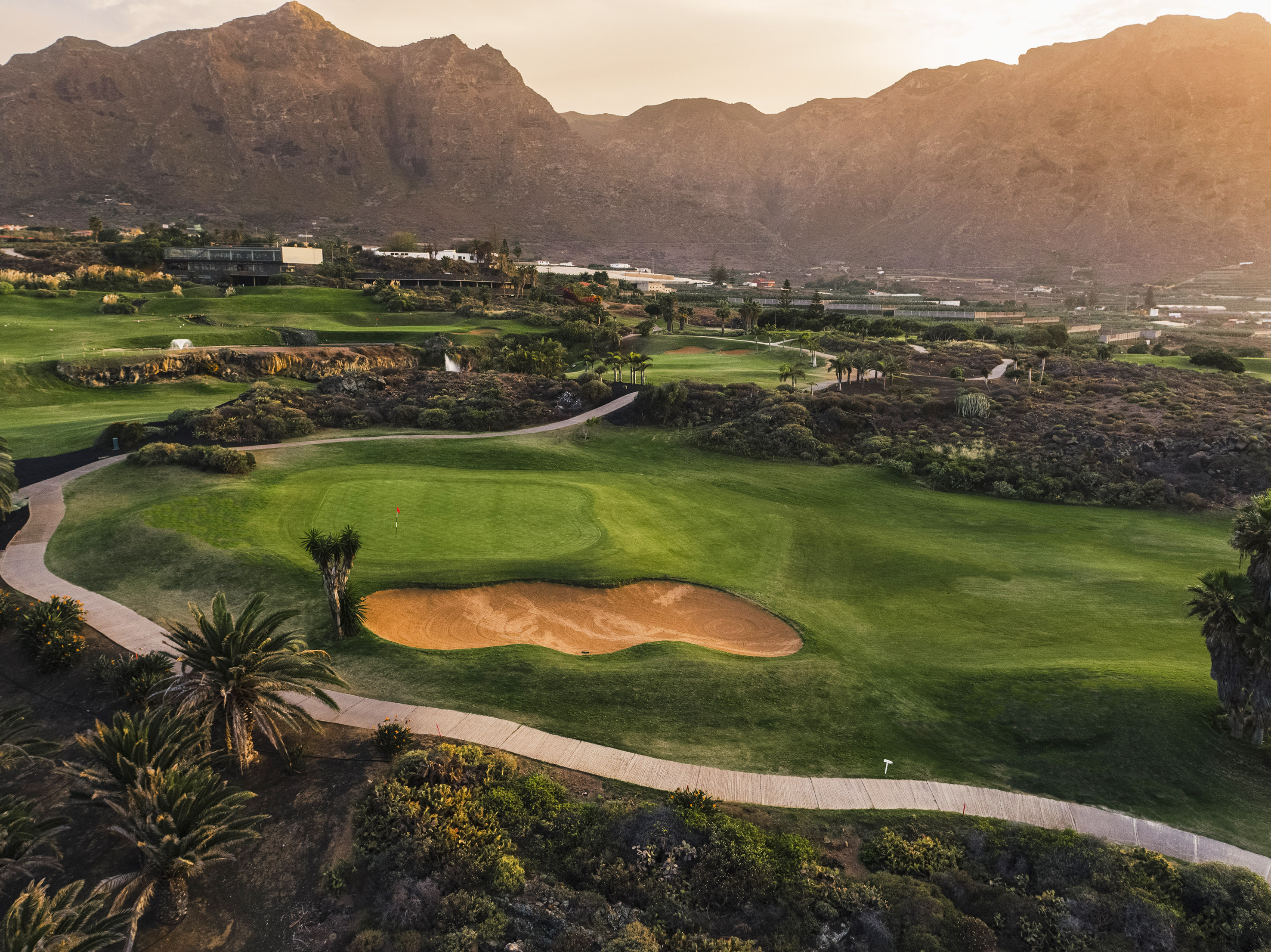 Mountain golf course with green fairways, sand bunker, palm trees, at sunset.