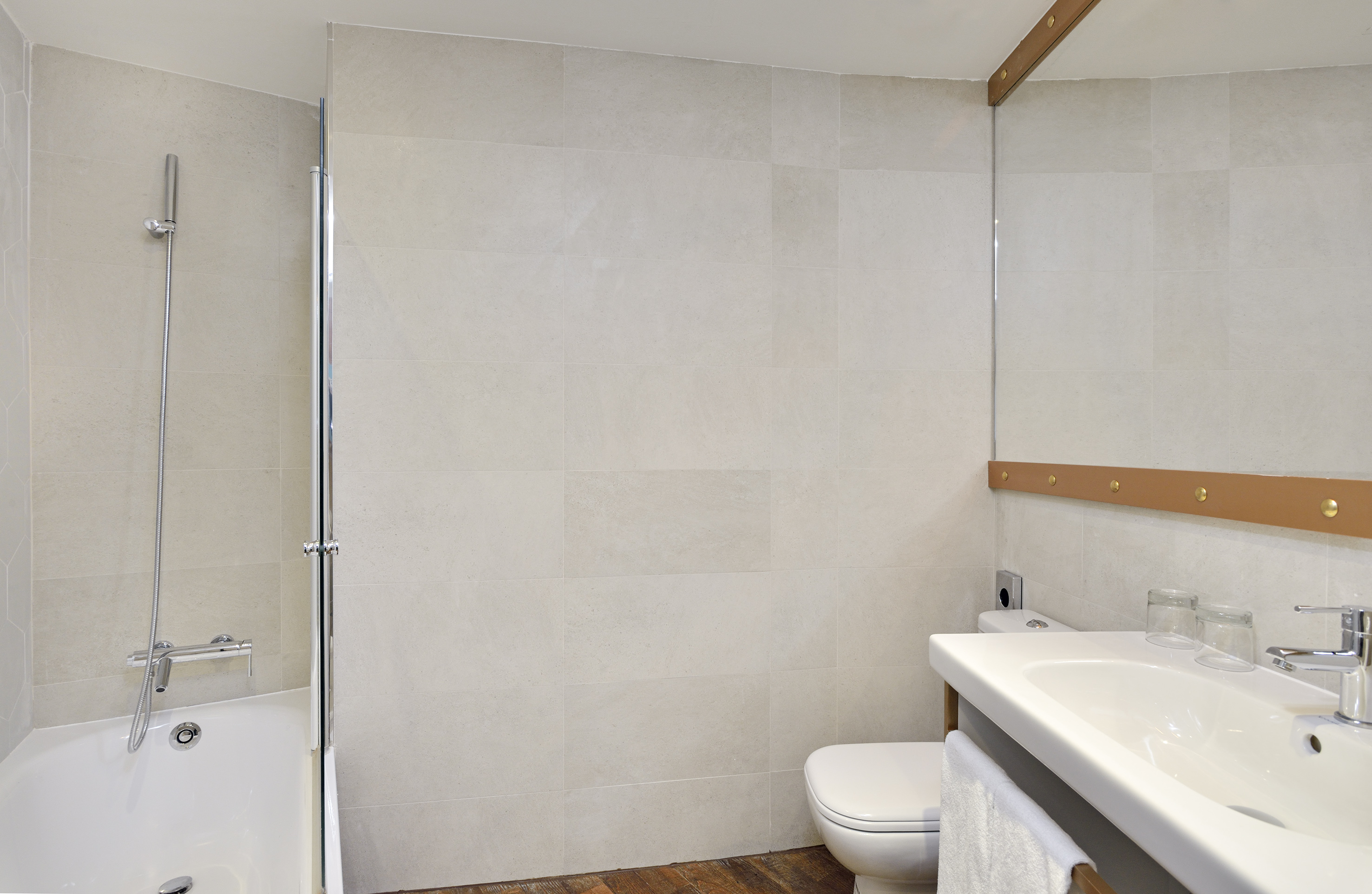 Minimalist hotel bathroom: white bathtub, shower, sink, toilet, light gray tiles.
