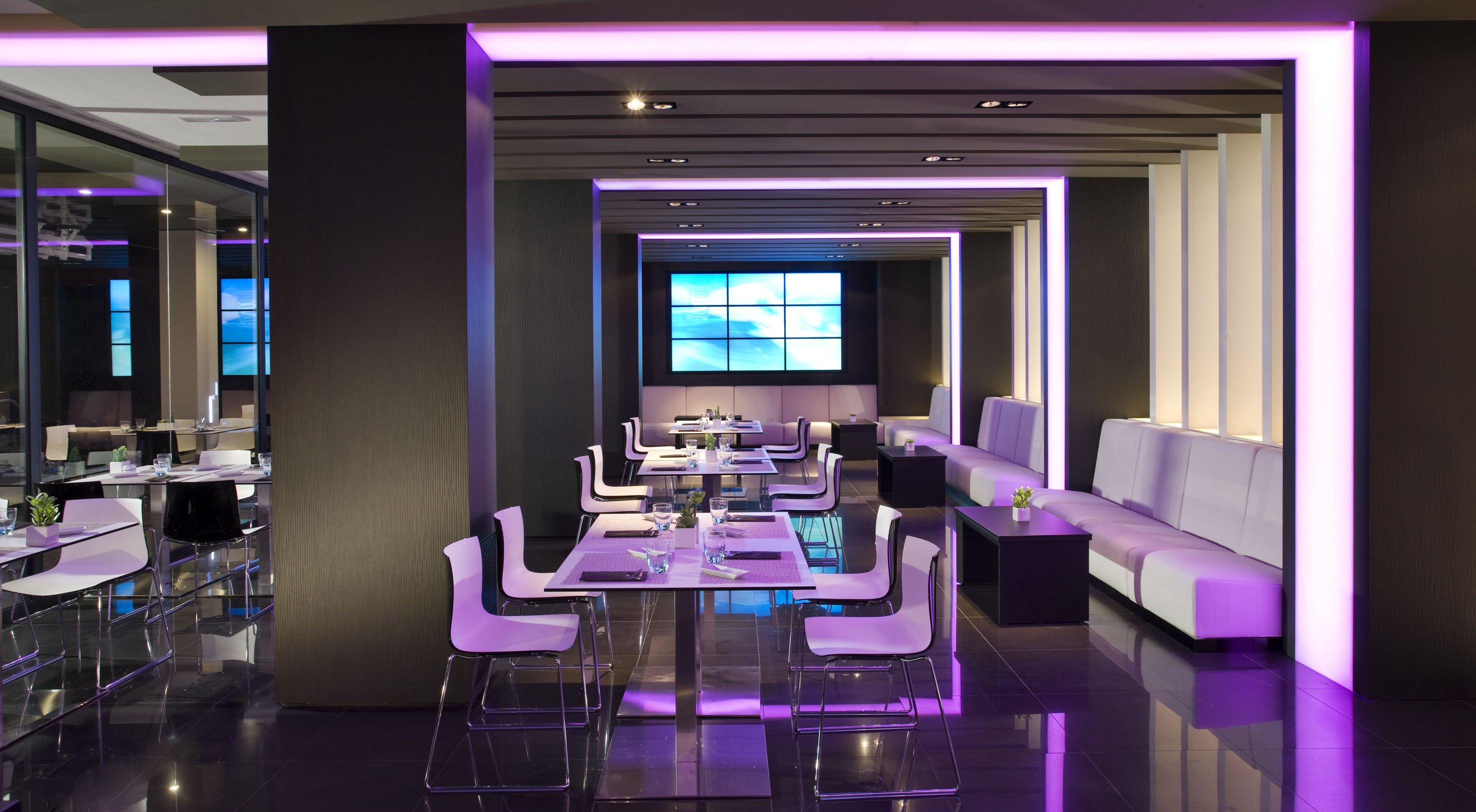 Modern lounge with purple lighting, sleek white seating, and large media screen.