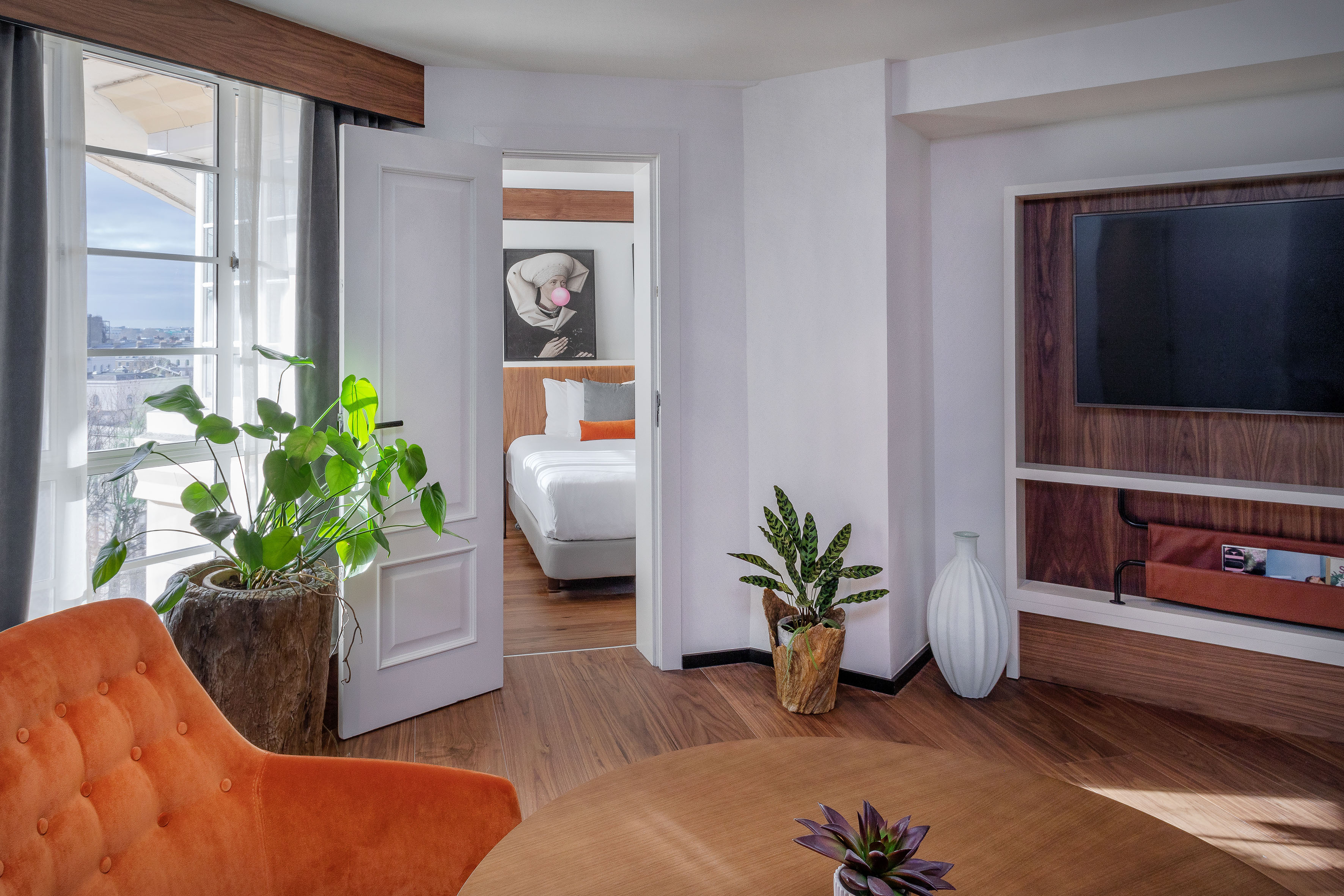 Chic hotel suite with orange armchair, TV, bedroom, and city view from window.