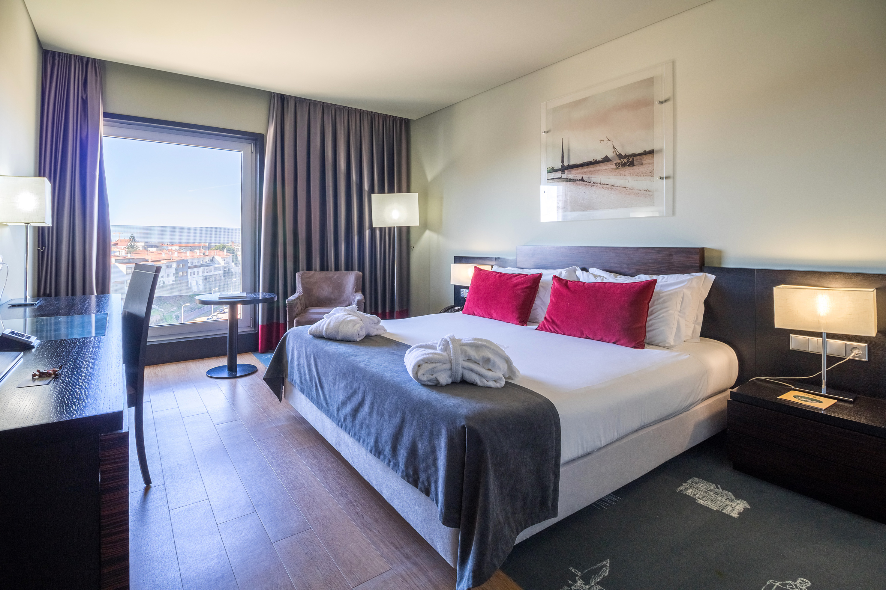 Modern hotel room with bed, red pillows, city view, desk, and bathrobes.