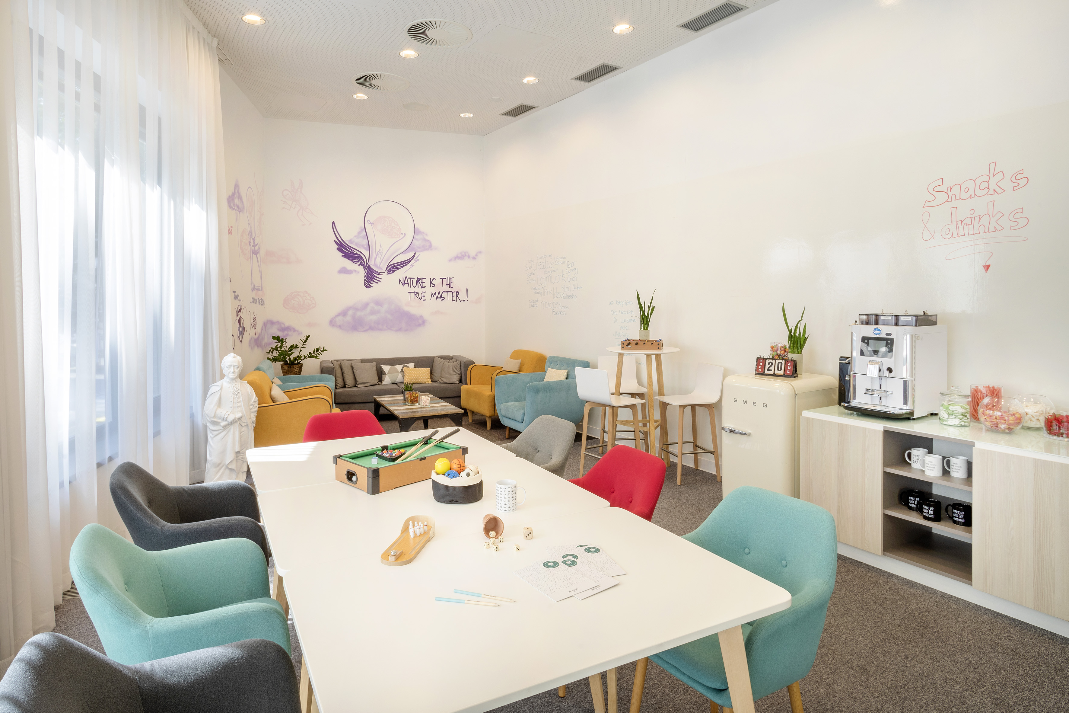 Modern office lounge with games table, colorful seating, coffee, and snacks.