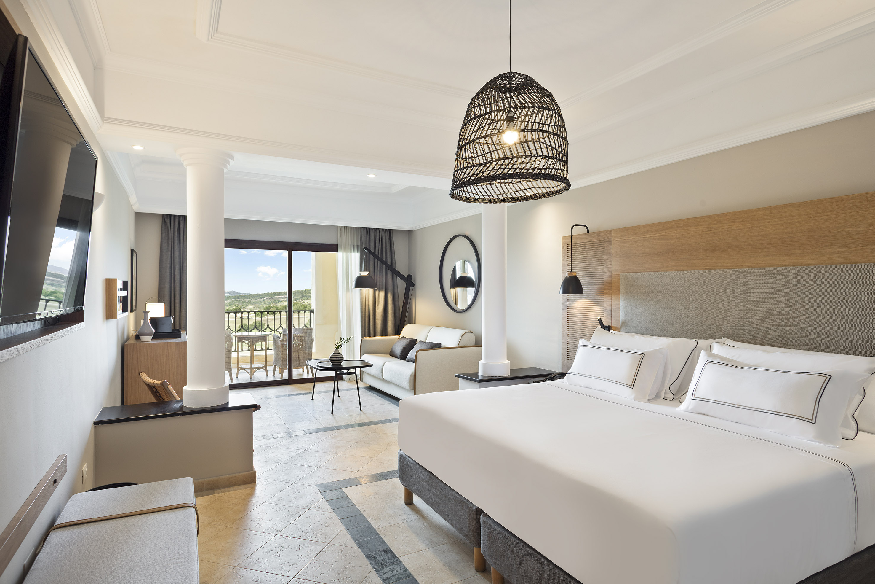 Modern hotel suite with king bed, sofa, balcony, and scenic landscape view.