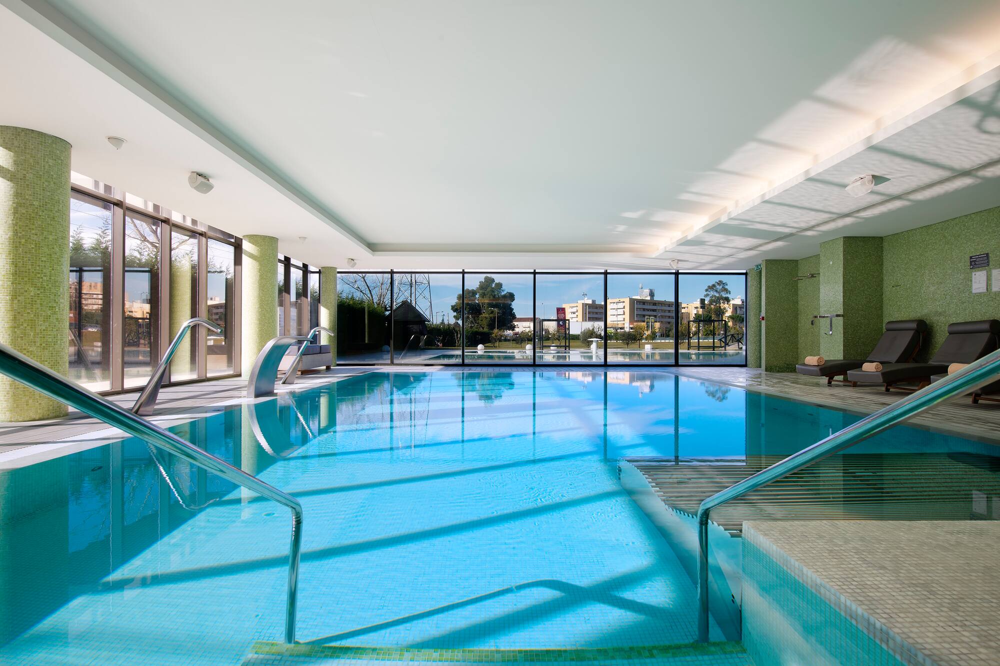 Modern indoor swimming pool with large windows, blue water, and green mosaic.