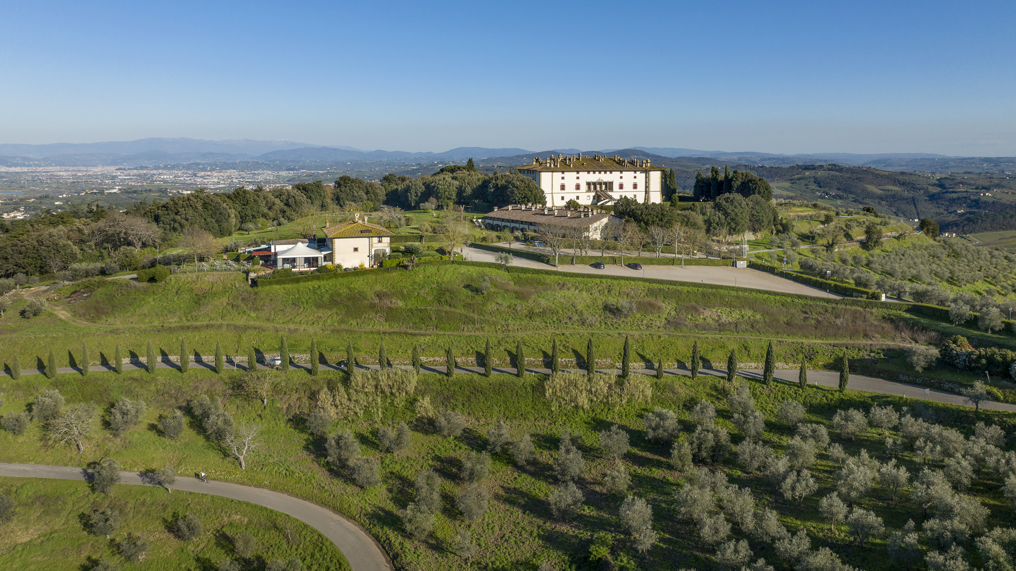 Grand Tuscan villa atop a green hill, with olive groves and cypress trees.