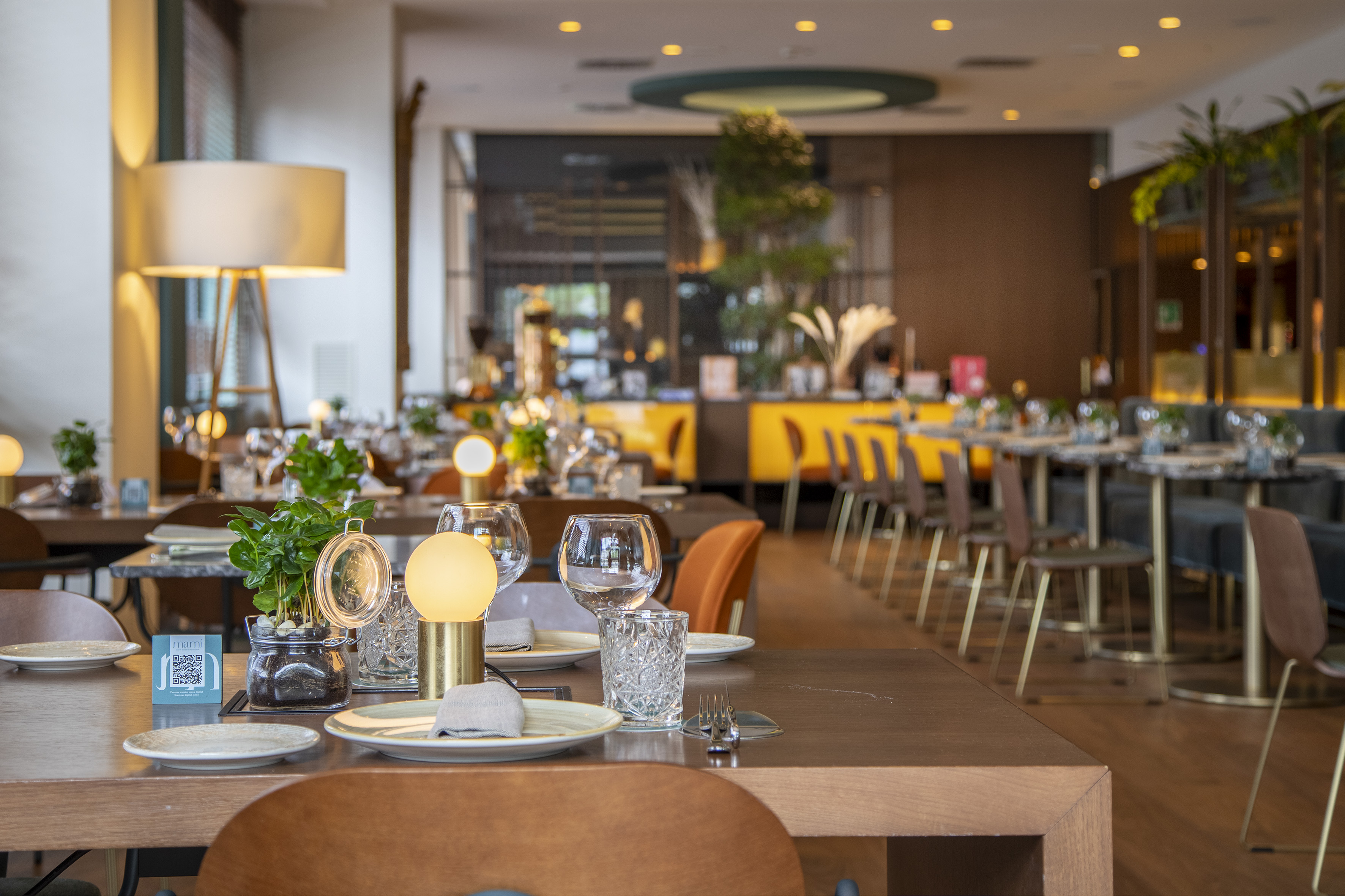 Modern restaurant dining room with set tables, plants, and warm lighting.