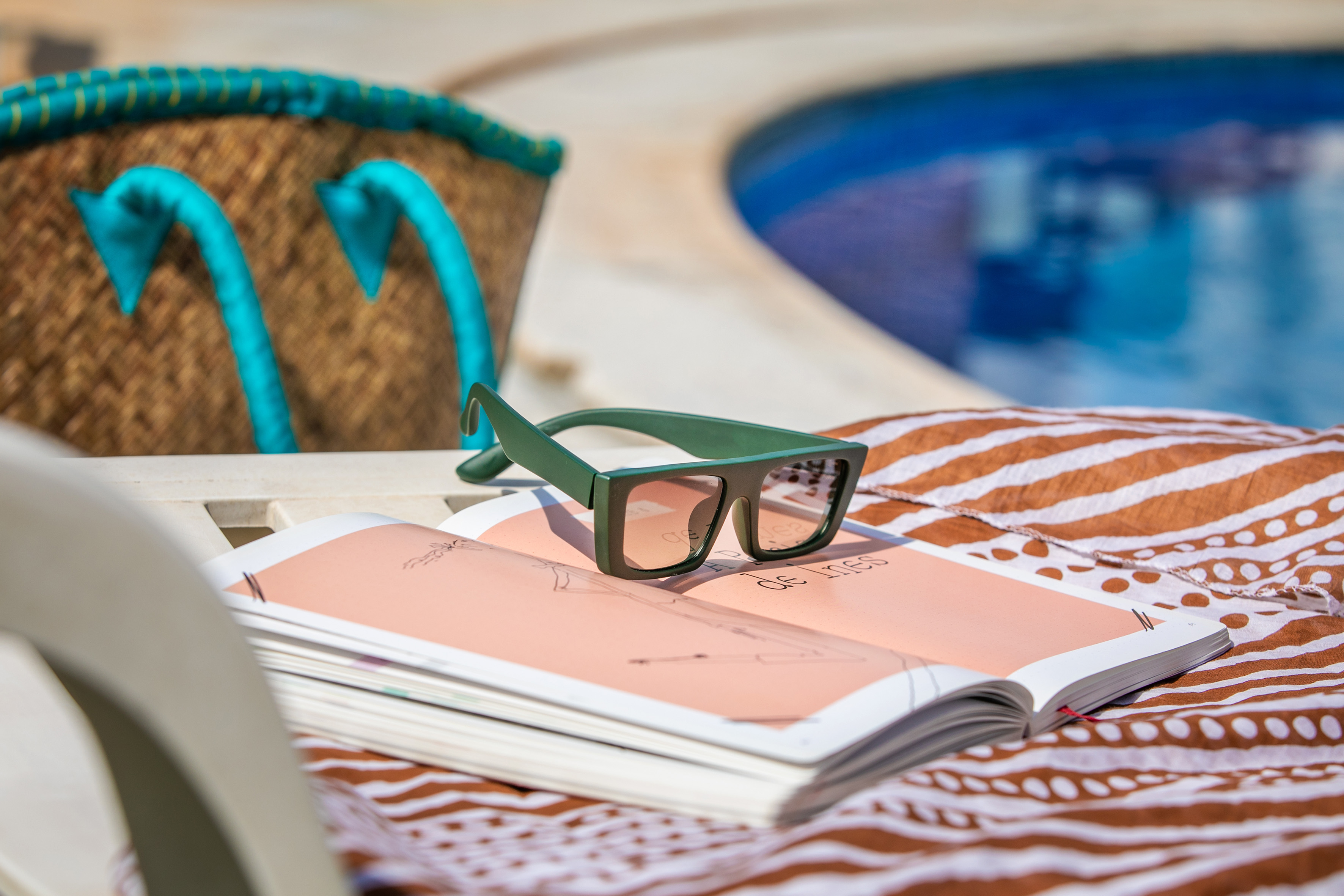 Green sunglasses on an open book, poolside with a blue pool and beach bag.
