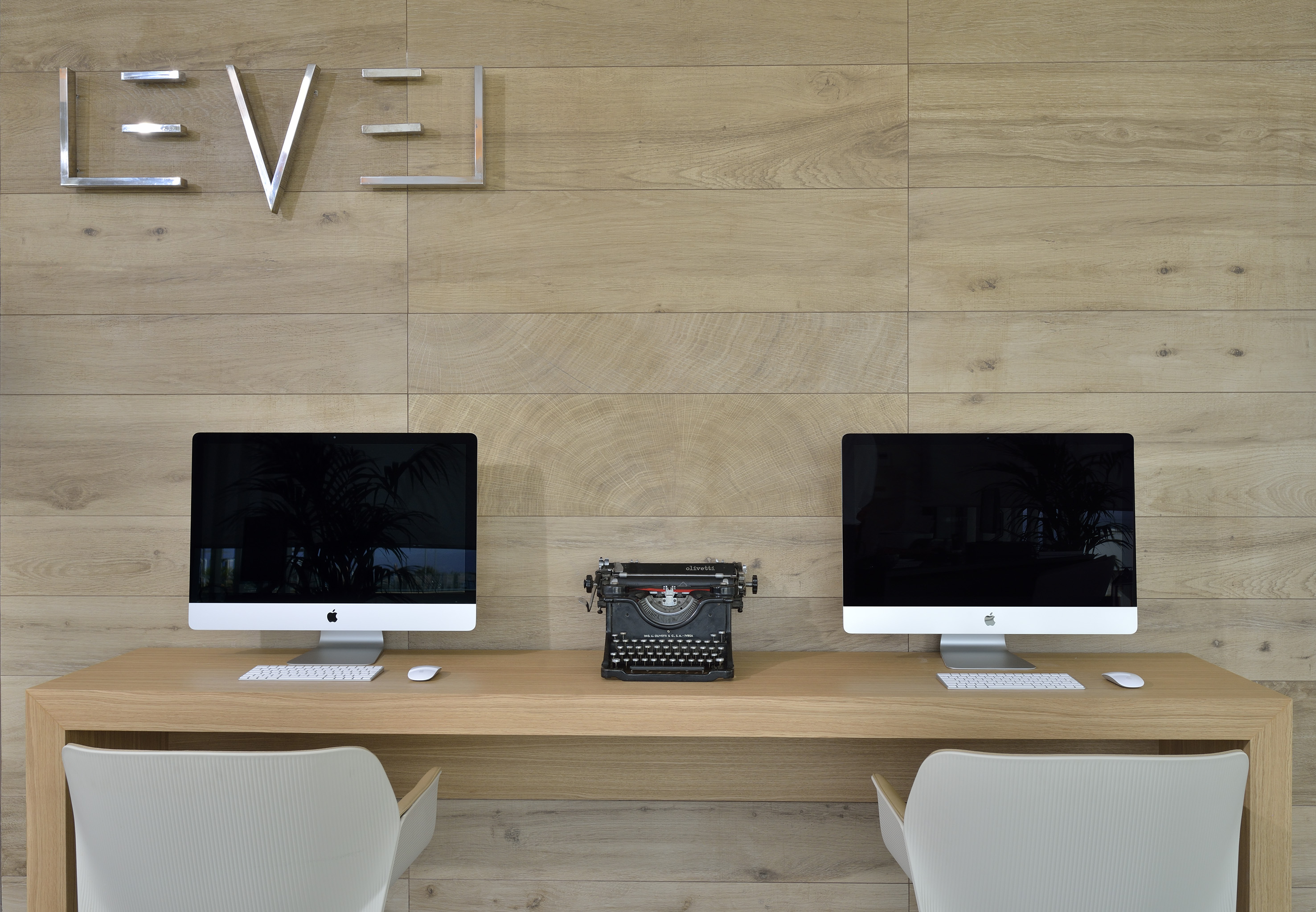 Modern office desk with dual iMacs, vintage typewriter, and 'LEVEL' wall sign.