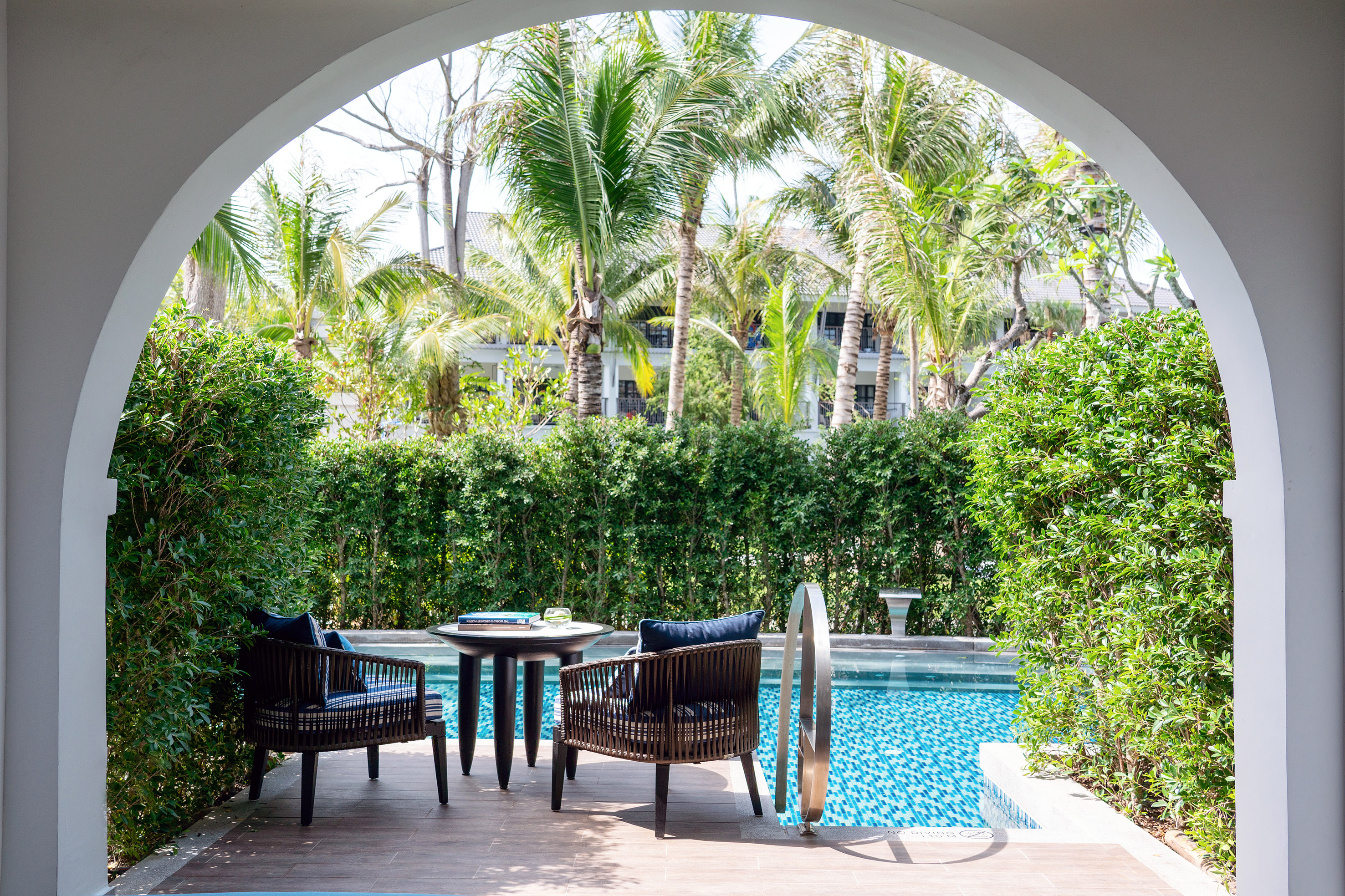 Tropical private pool patio with outdoor seating, hedges, and palm trees.