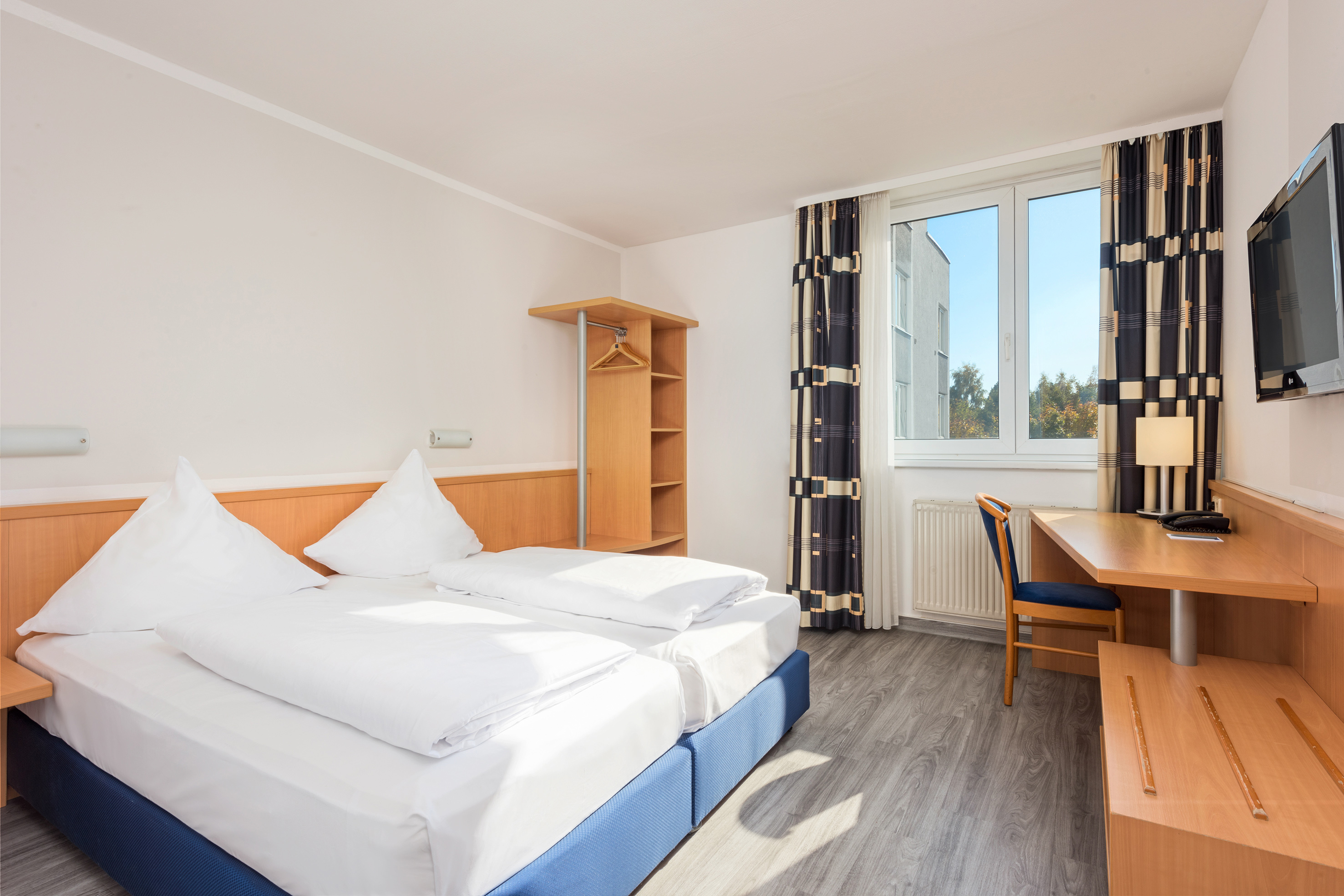 Bright hotel room with twin beds, wooden desk, TV, and large window.