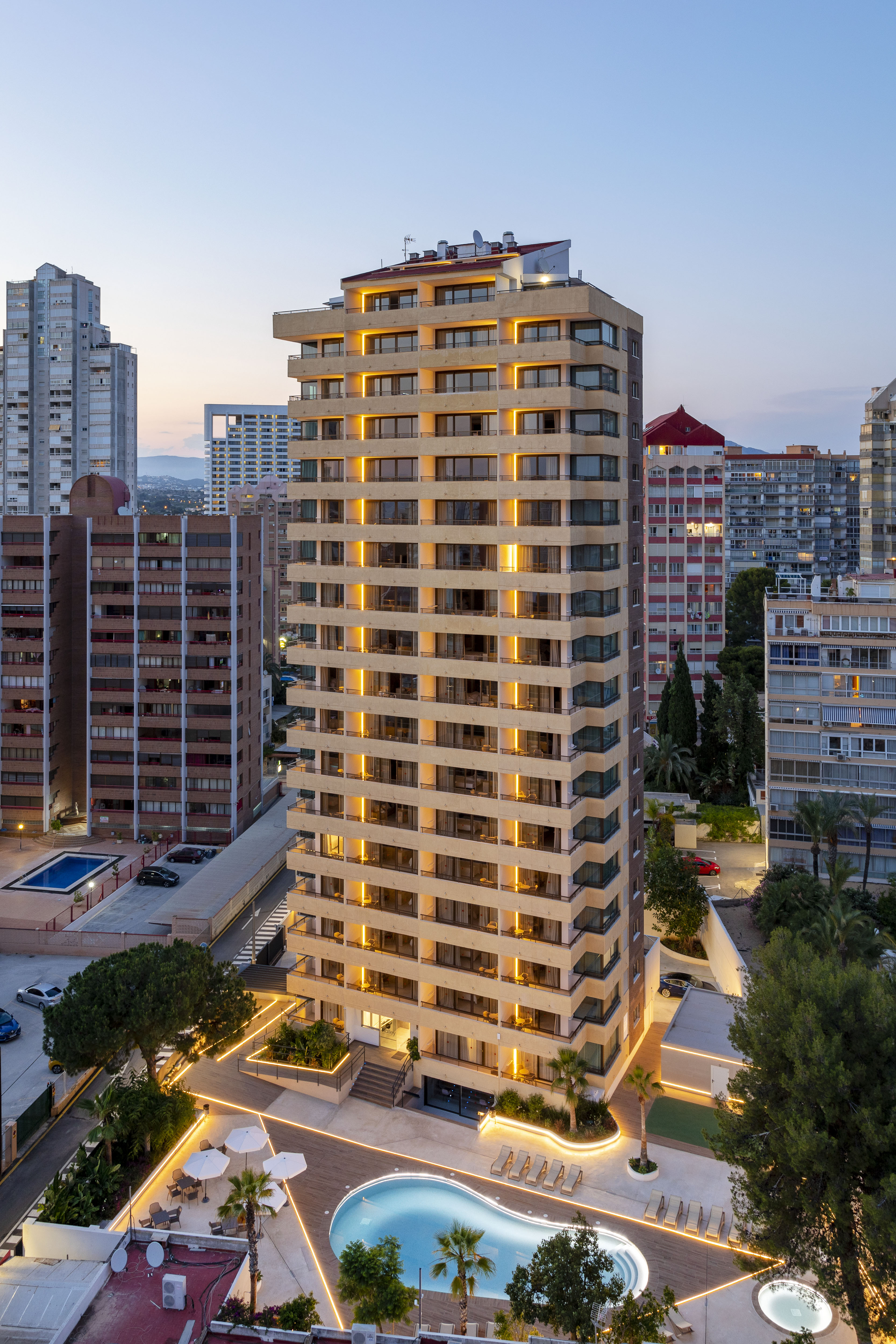 Tall illuminated building with outdoor swimming pool at dusk, urban cityscape.