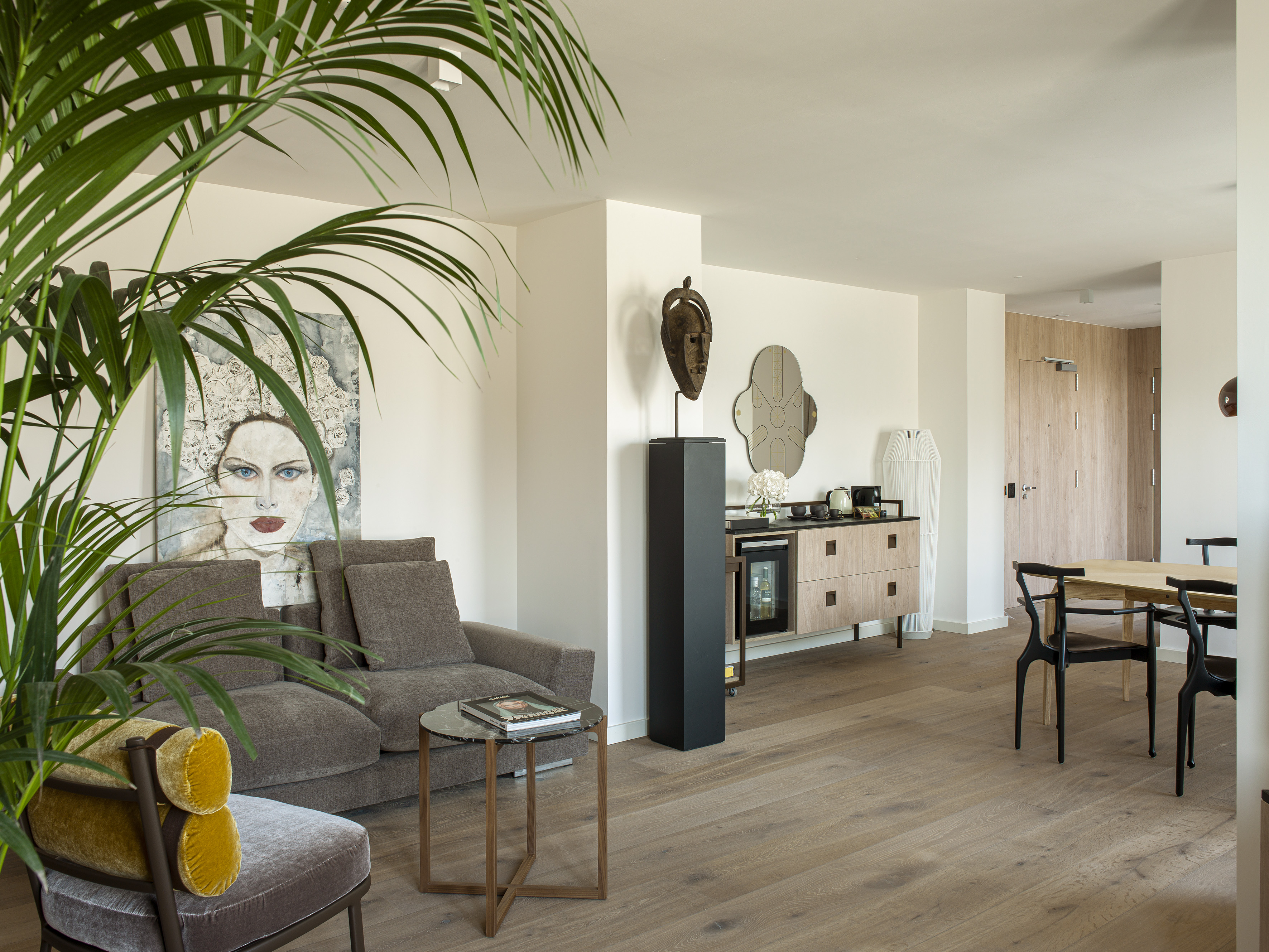 Modern apartment living room with gray sofa, palm plant, abstract art, dining table.