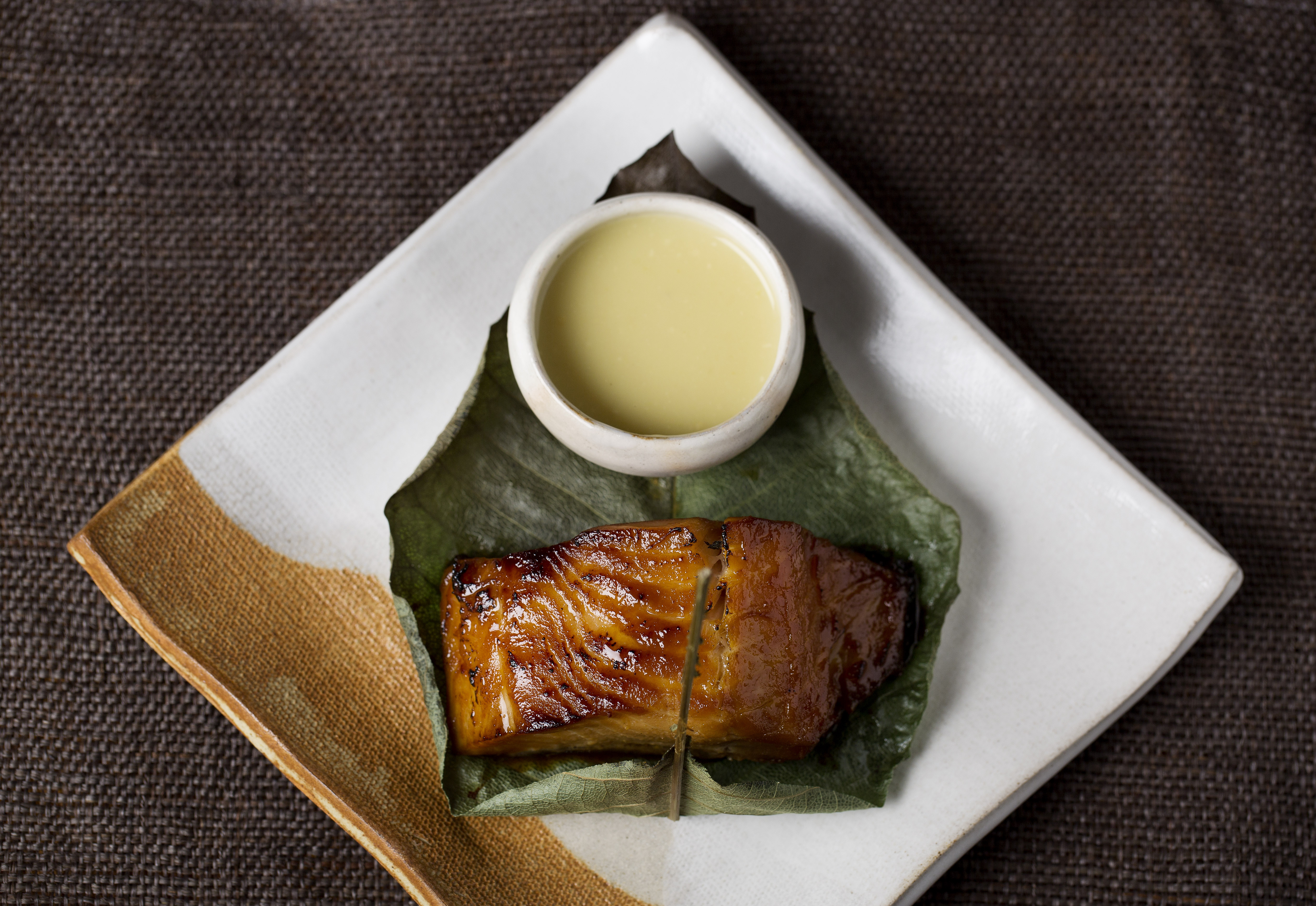 Miso glazed black cod with pale yellow sauce on a leaf, white plate.