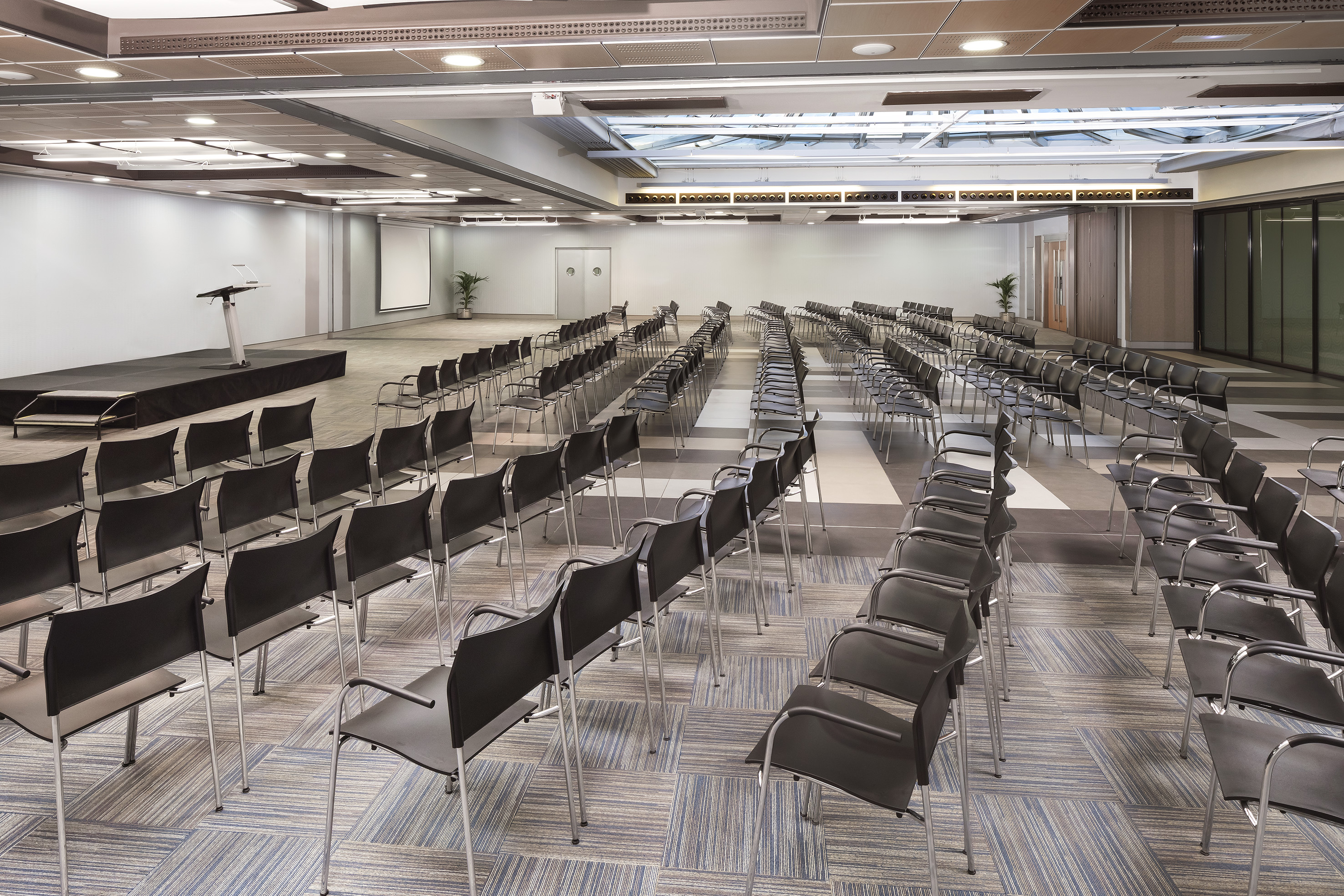 Empty modern conference room with stage, lectern, screen, and rows of black chairs.