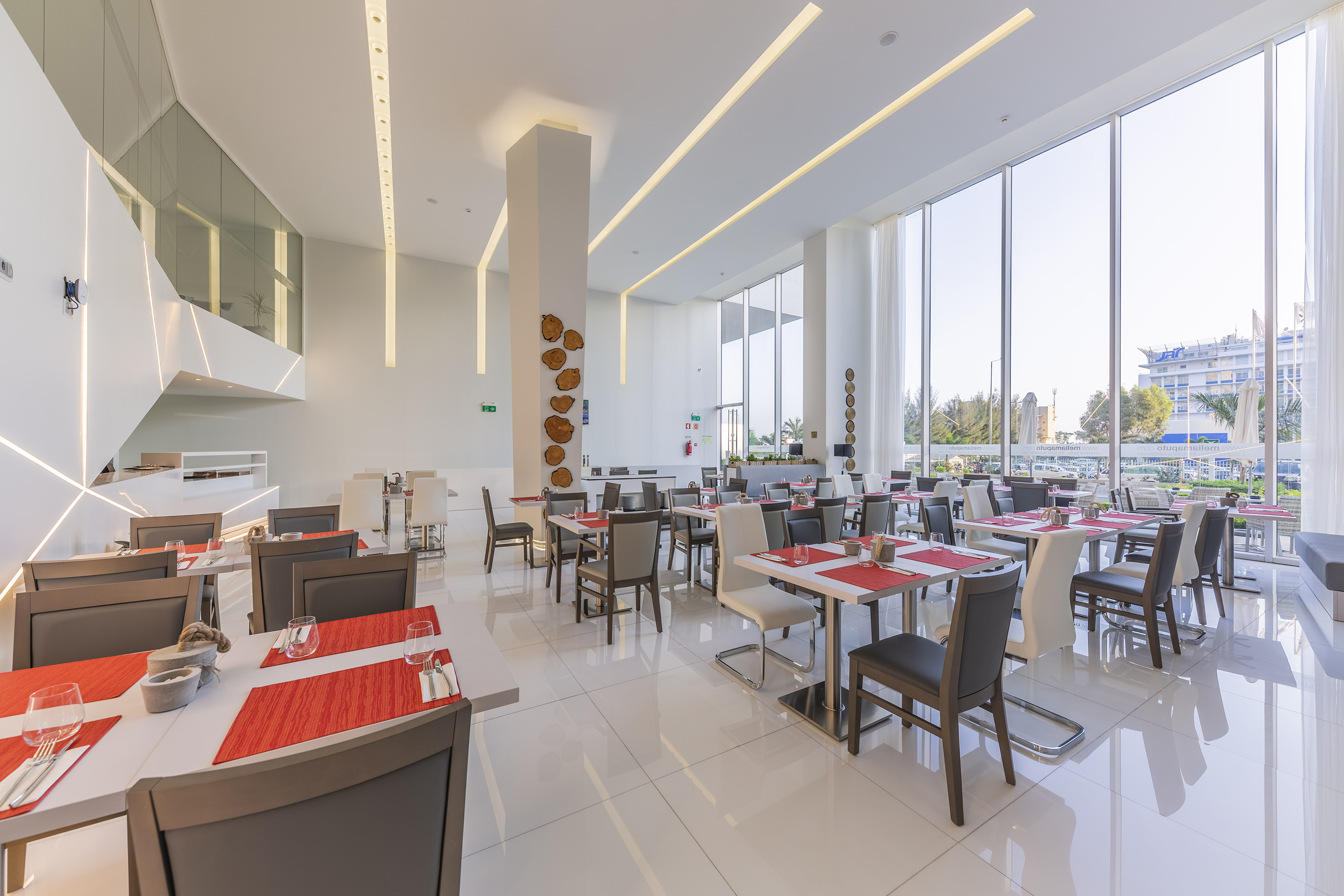 Modern hotel restaurant with large windows, white decor, and red table settings. - Koffiebar (Bar), Aperitieven - Meliá Maputo Sky, Maputo