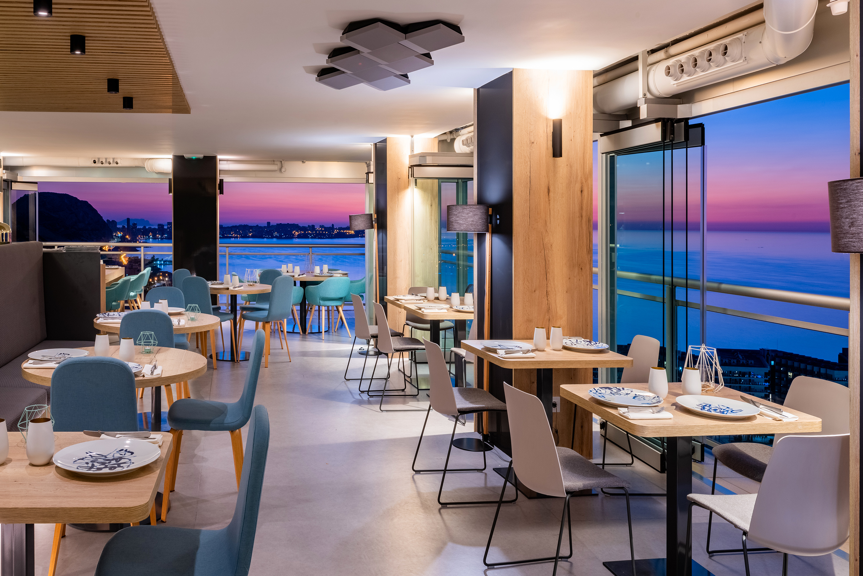 Modern restaurant dining room with ocean and city view at sunset.
