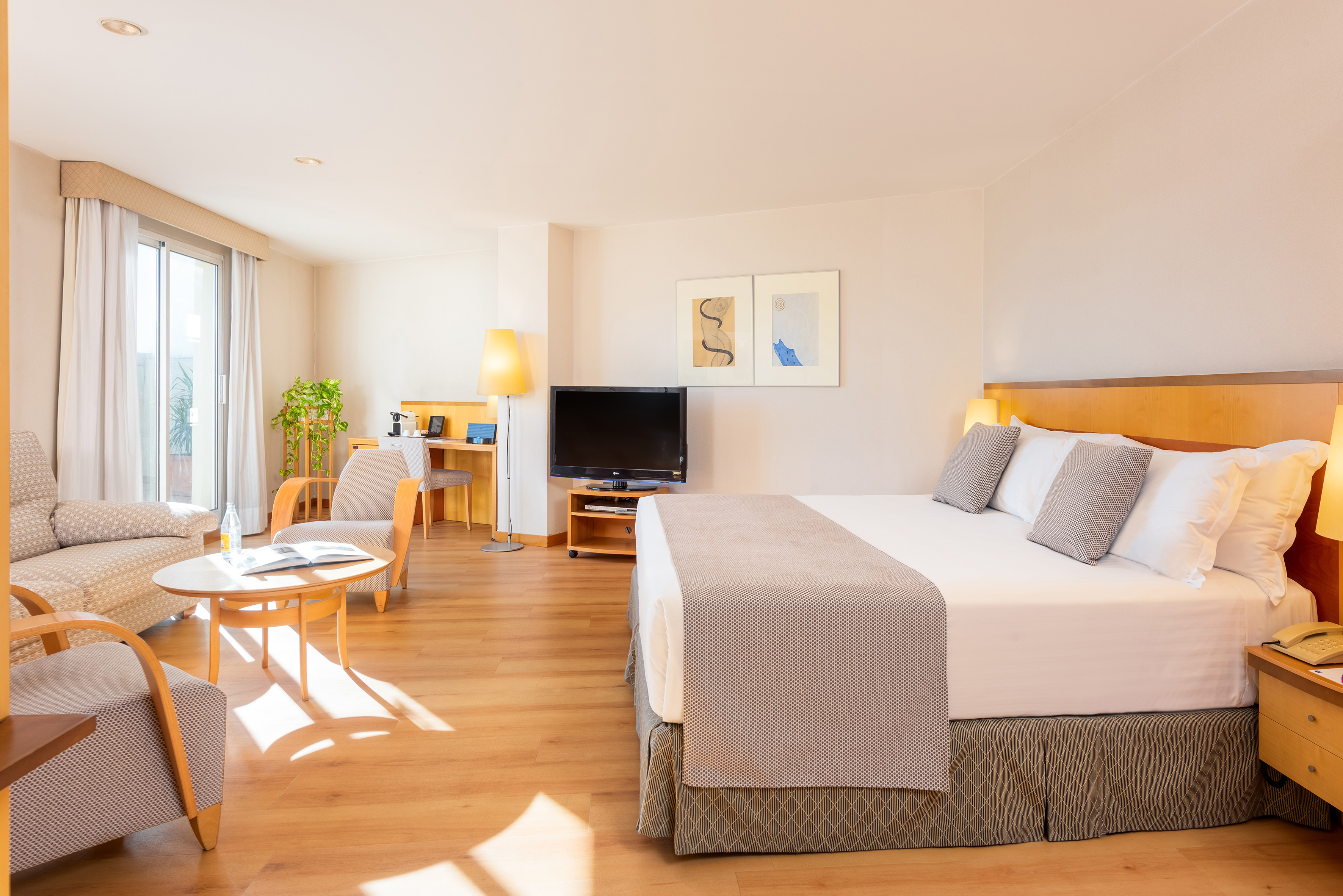 Bright hotel suite with large bed, sitting area, desk, TV, and wood floor.