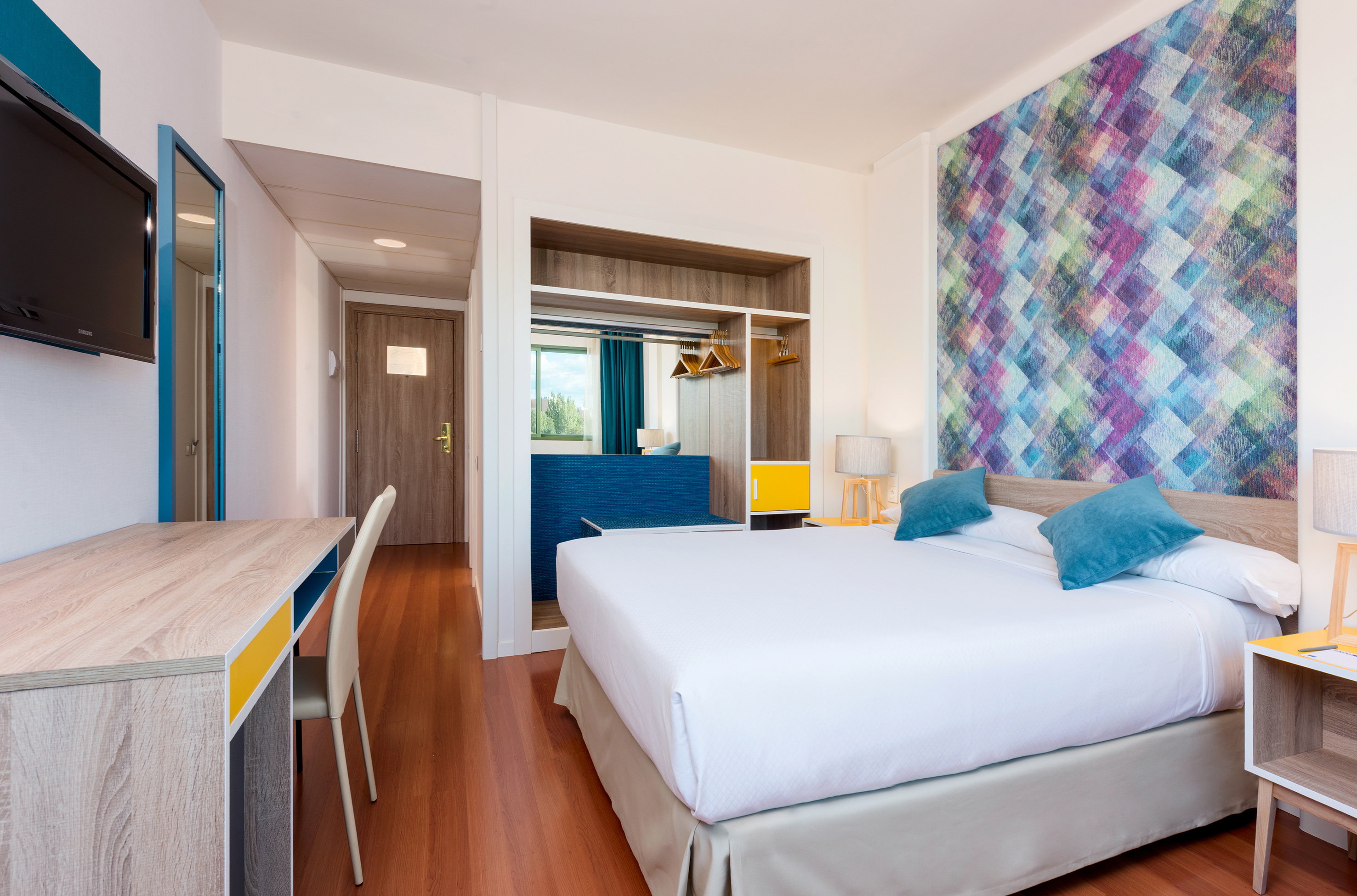 Modern hotel room with king bed, colorful geometric wall, desk, and open closet.