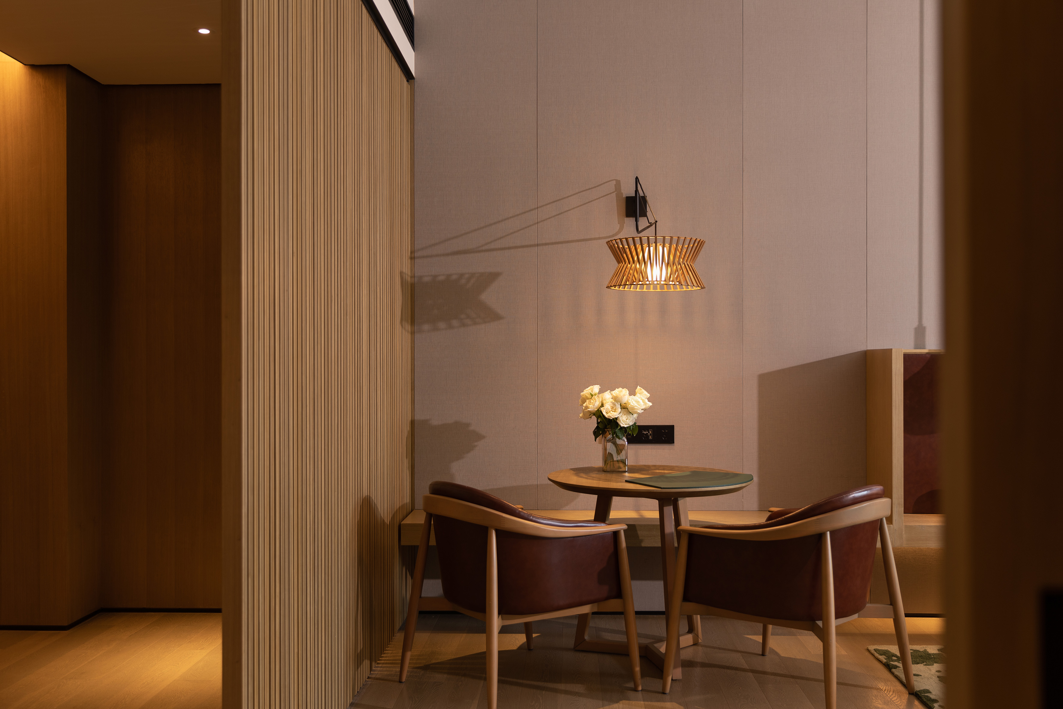 Modern hotel nook with wooden table, leather chairs, and slatted wall lamp.