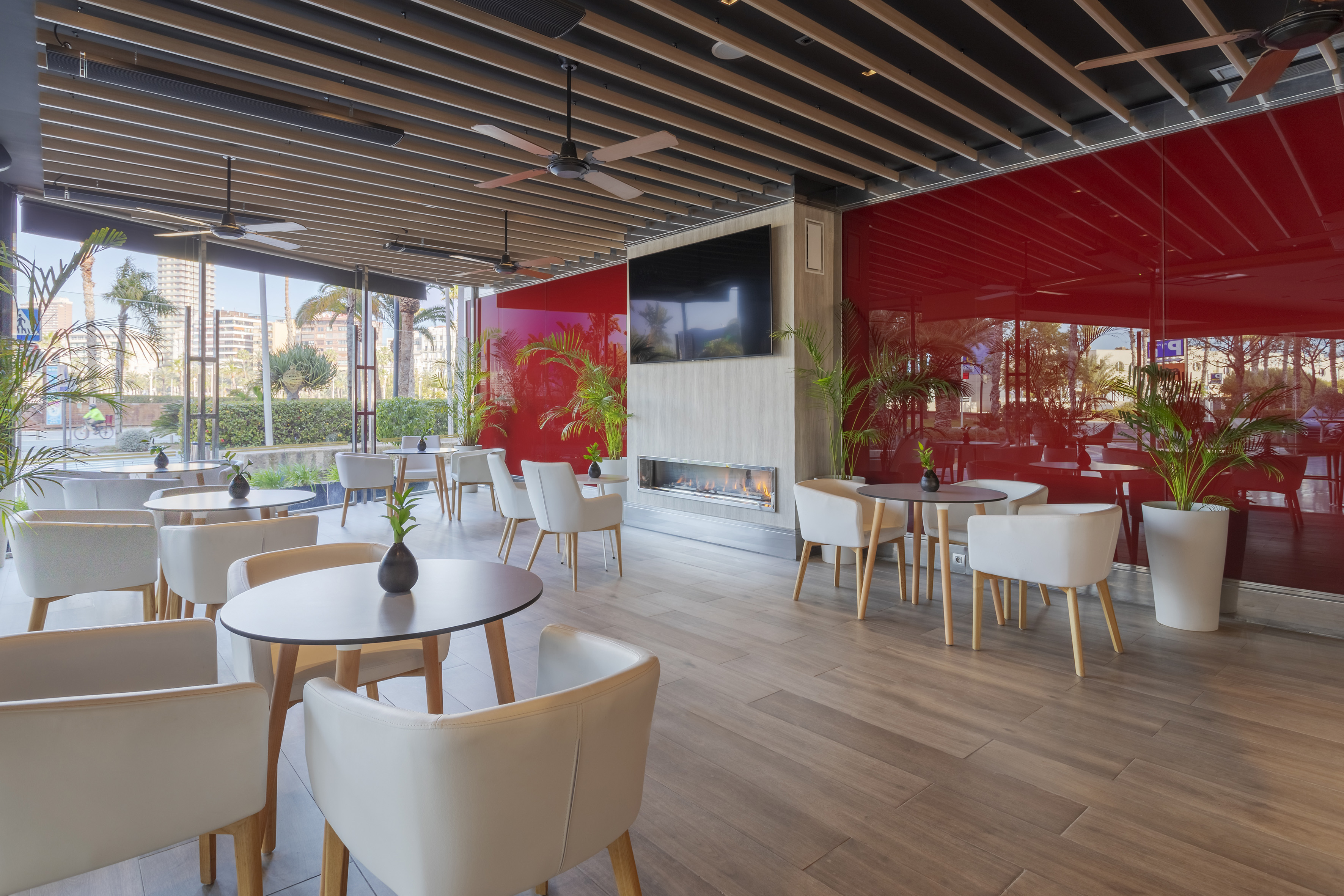 Modern restaurant with red accent wall, fireplace, white seating, city view.