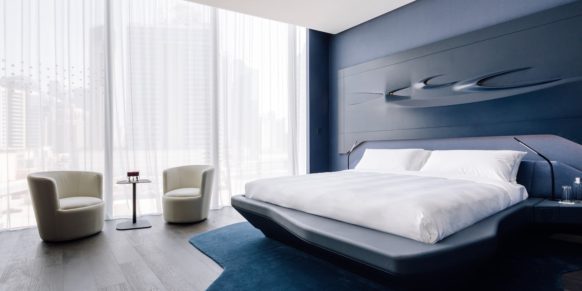 Modern blue bedroom with sculptural headboard, city view, and contemporary seating.